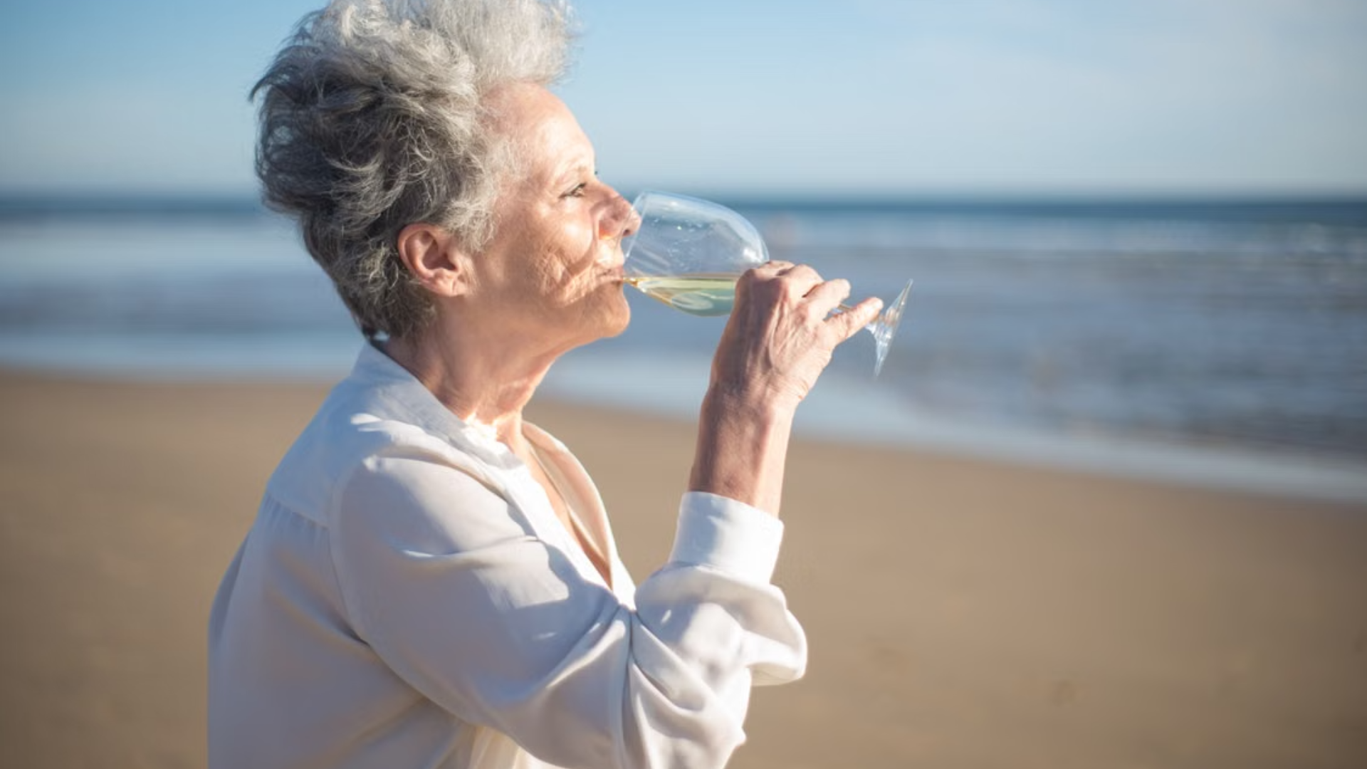 An older woman with grey hair enjoys a glass of white wine while relaxing on a sunny beach.
