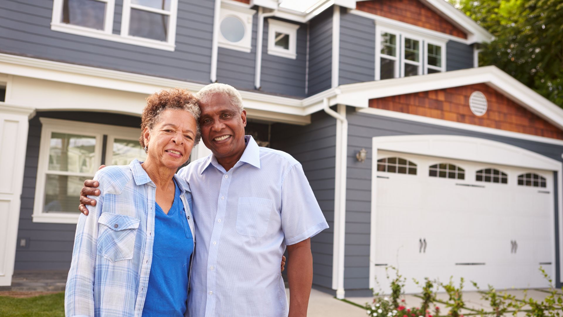 Smiling older couple embracing in front of a large suburban house.