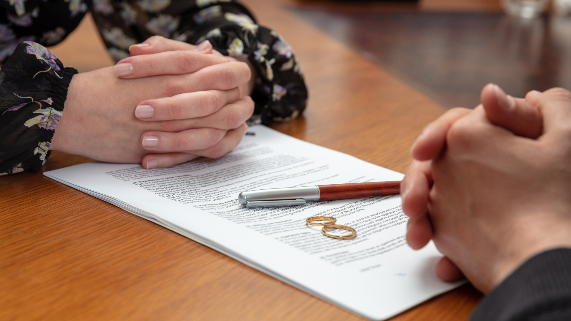 A close-up view shows two people sitting across a wooden table with hands clasped over divorce documents and two gold wedding rings next to a pen.