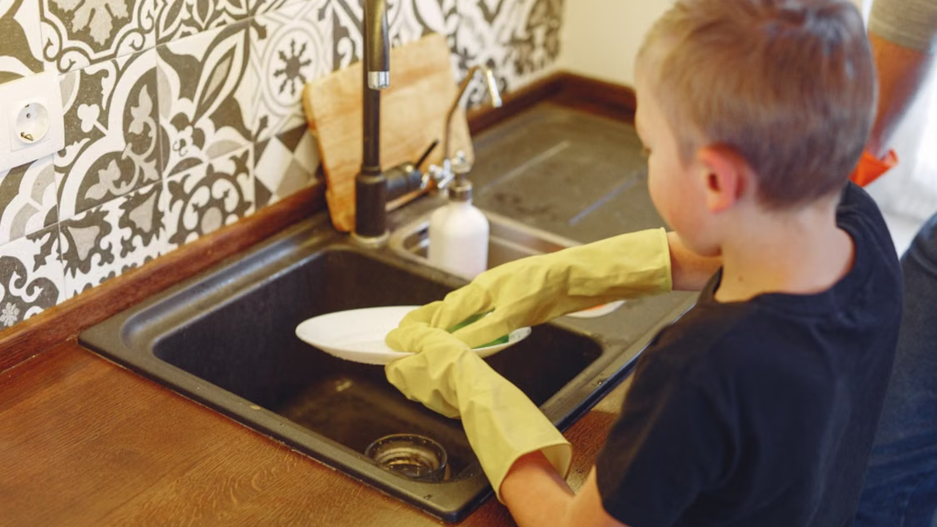 A young boy wearing oversized yellow rubber gloves washes a white plate in a kitchen sink.