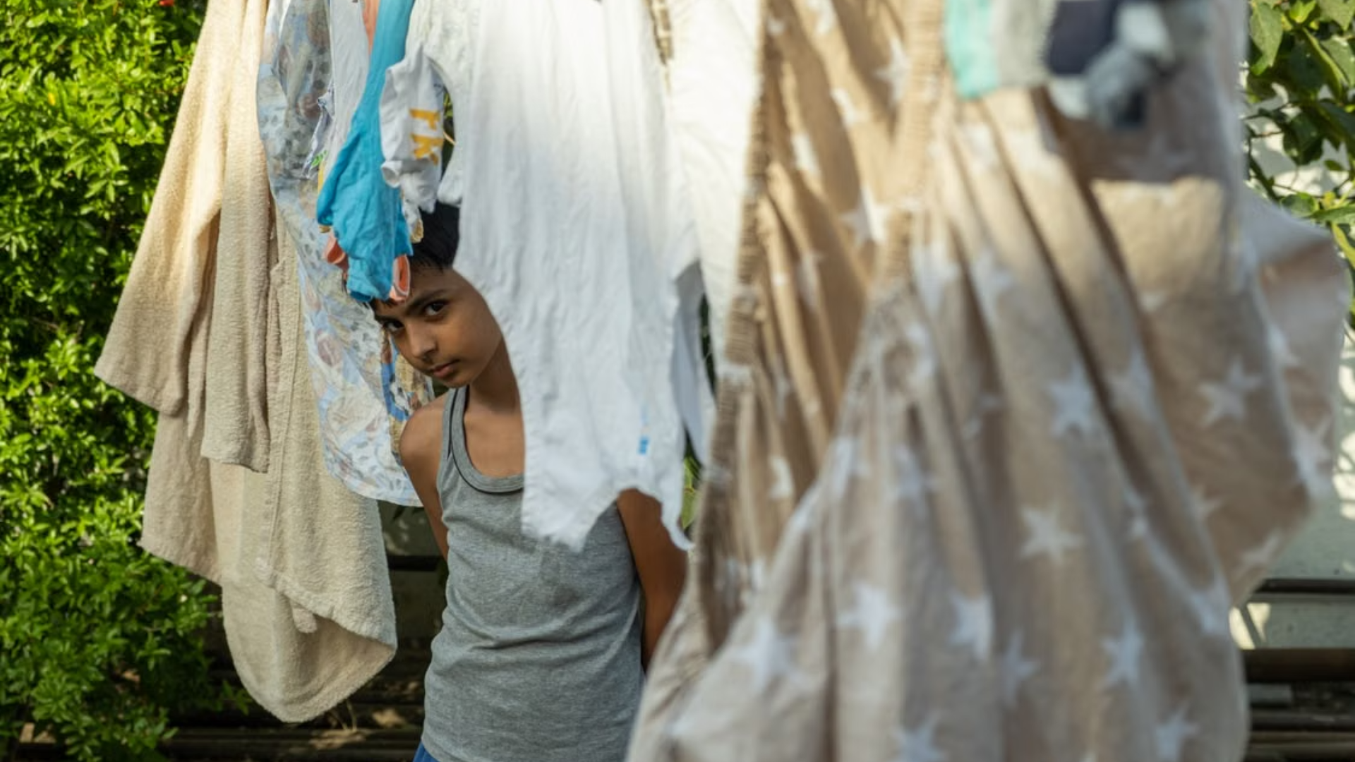 A young boy peers from behind clothing and bedding hanging on a clothesline outdoors.