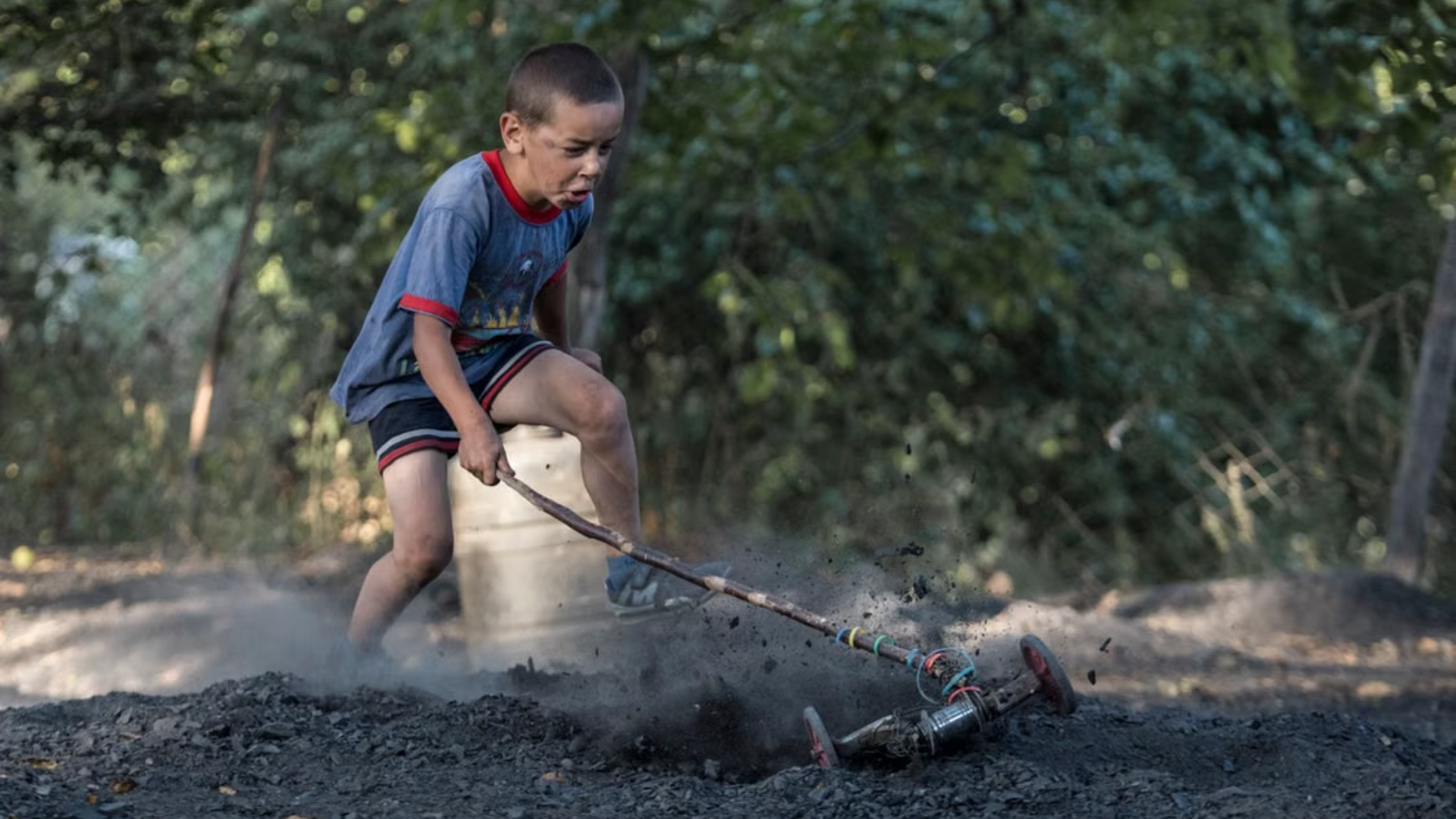 A young boy plays outdoors, aggressively pushing a makeshift lawn mower.