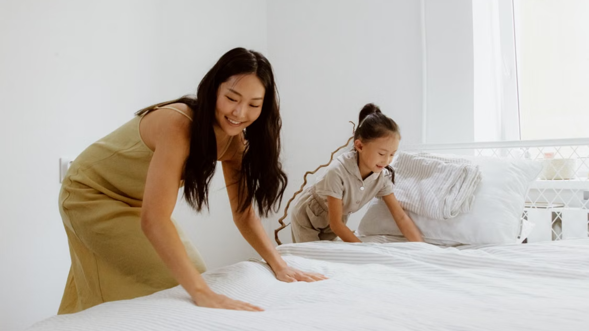 A smiling woman and a young girl work together to make a bed in a bright, white bedroom.