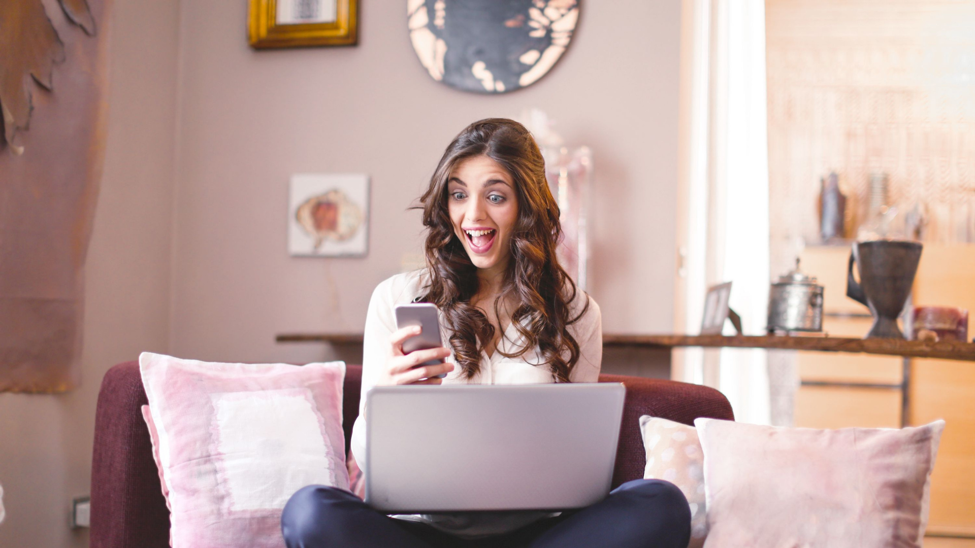 Smiling woman at home, using smartphone and laptop, expressing surprise and happiness.