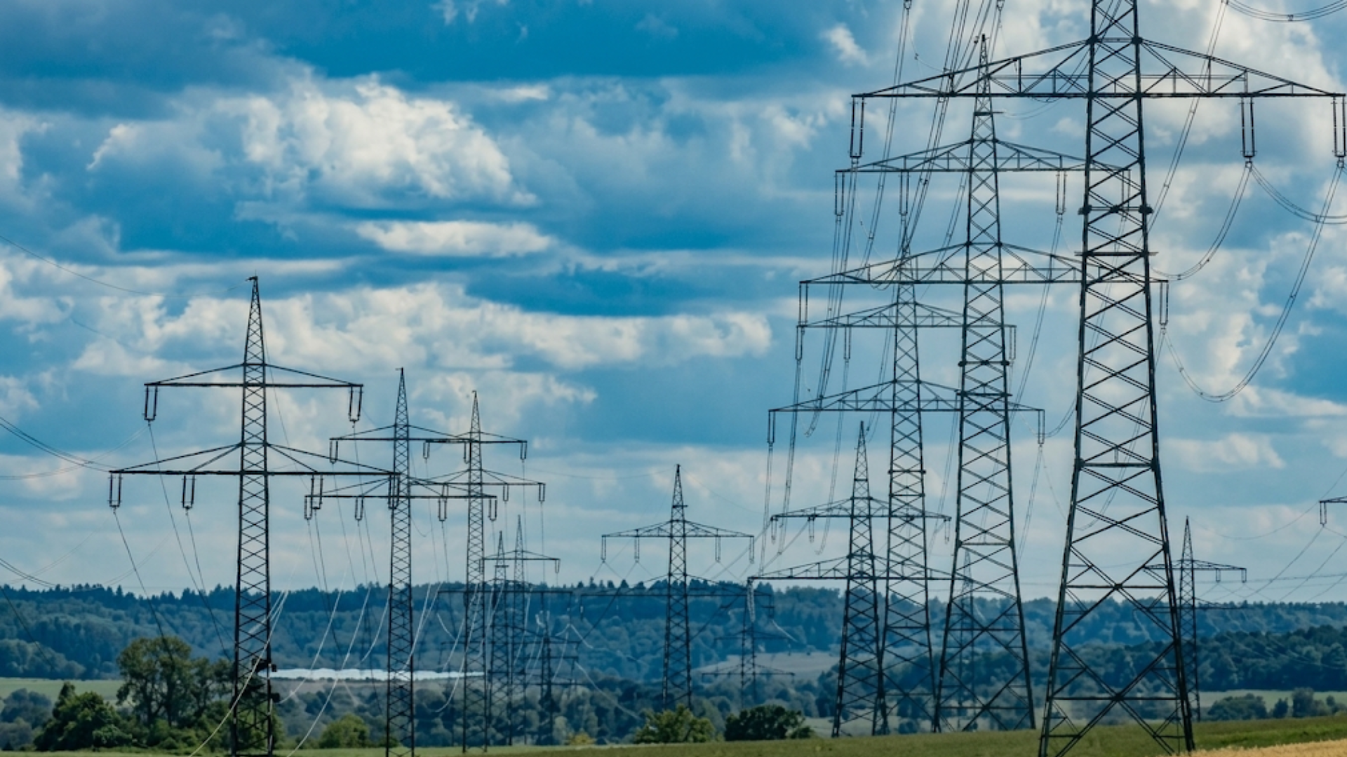 Multiple high-voltage transmission towers and power lines across a grassy landscape under a cloudy sky.