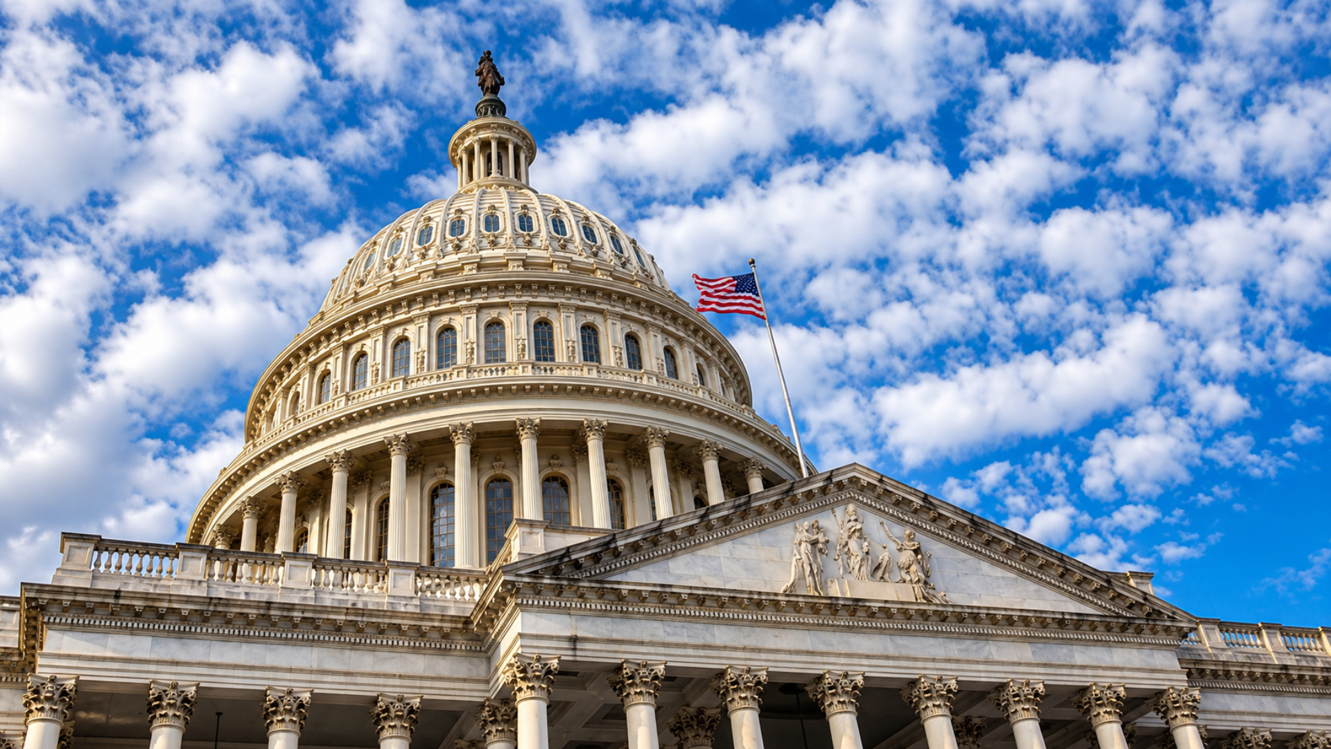 United States Capitol dome and pediment featuring the American flag under scattered clouds.