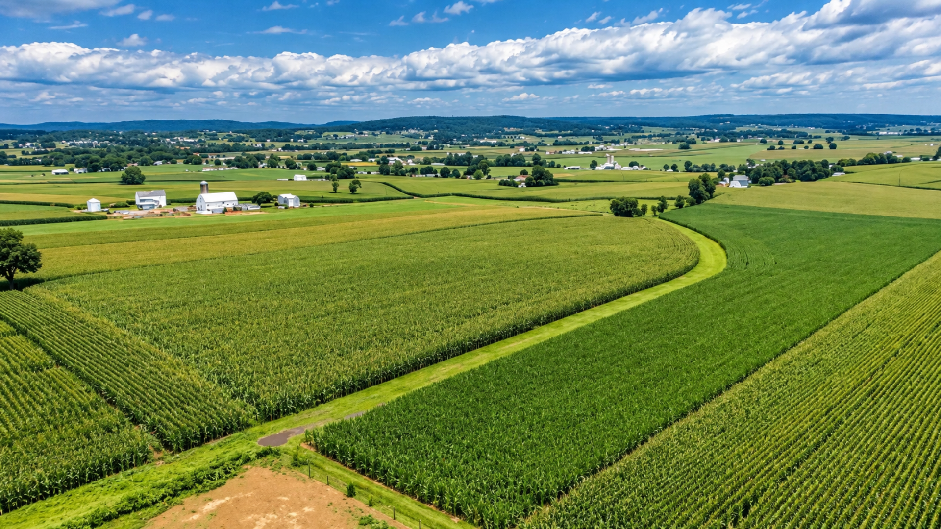 High-angle landscape view of green agricultural crop fields and farm structures under a blue sky.