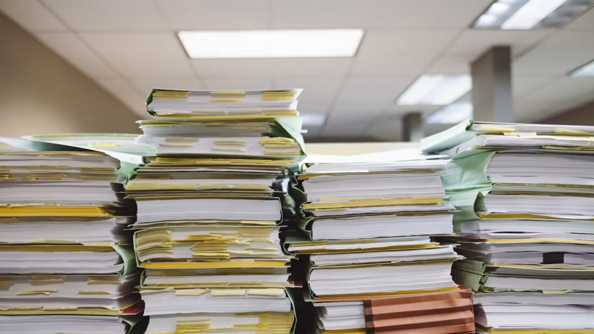 Tall stacks of paper documents and files organized with sticky notes on a desk.