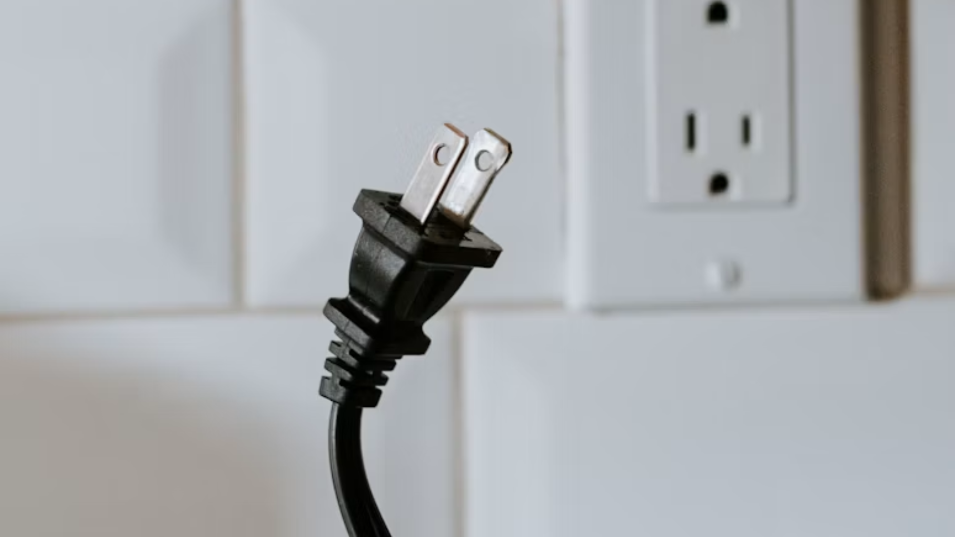 Close-up of a two-prong black electrical plug held up against a tiled wall.