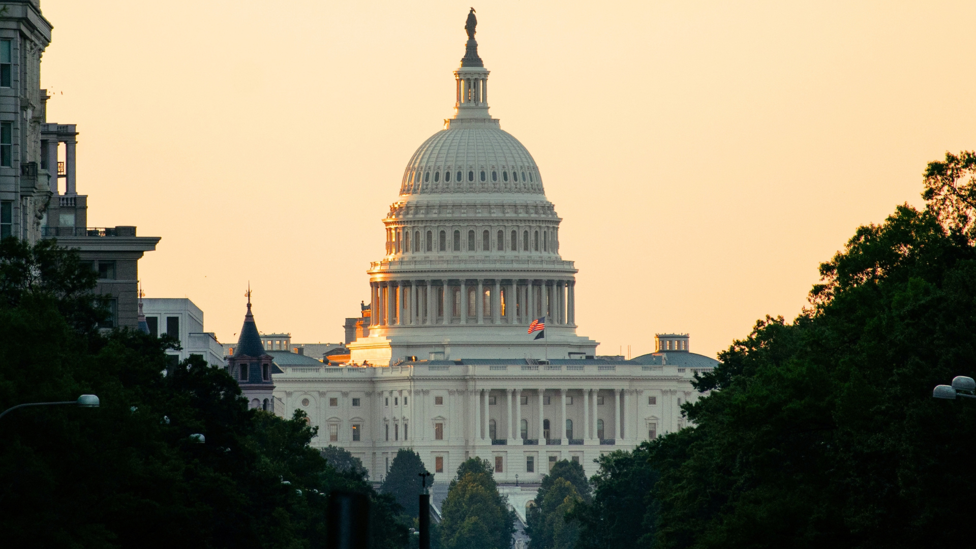 The United States Capitol building illuminated by the warm light of sunrise or sunset, framed by dark green trees in the foreground.