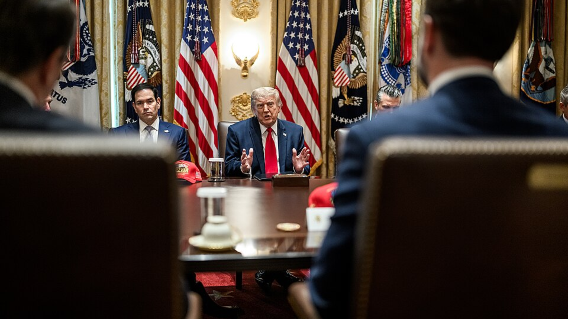 Donald Trump speaking at a conference table, flanked by Senator Marco Rubio and other officials, with US flags in the background.
