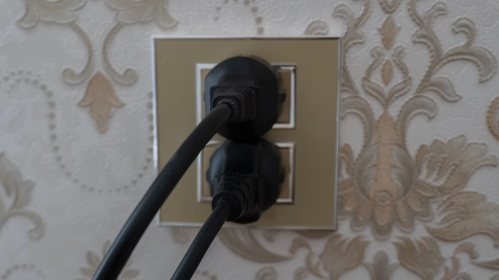 Two black electrical cords plugged into a dual wall outlet set against patterned wallpaper.