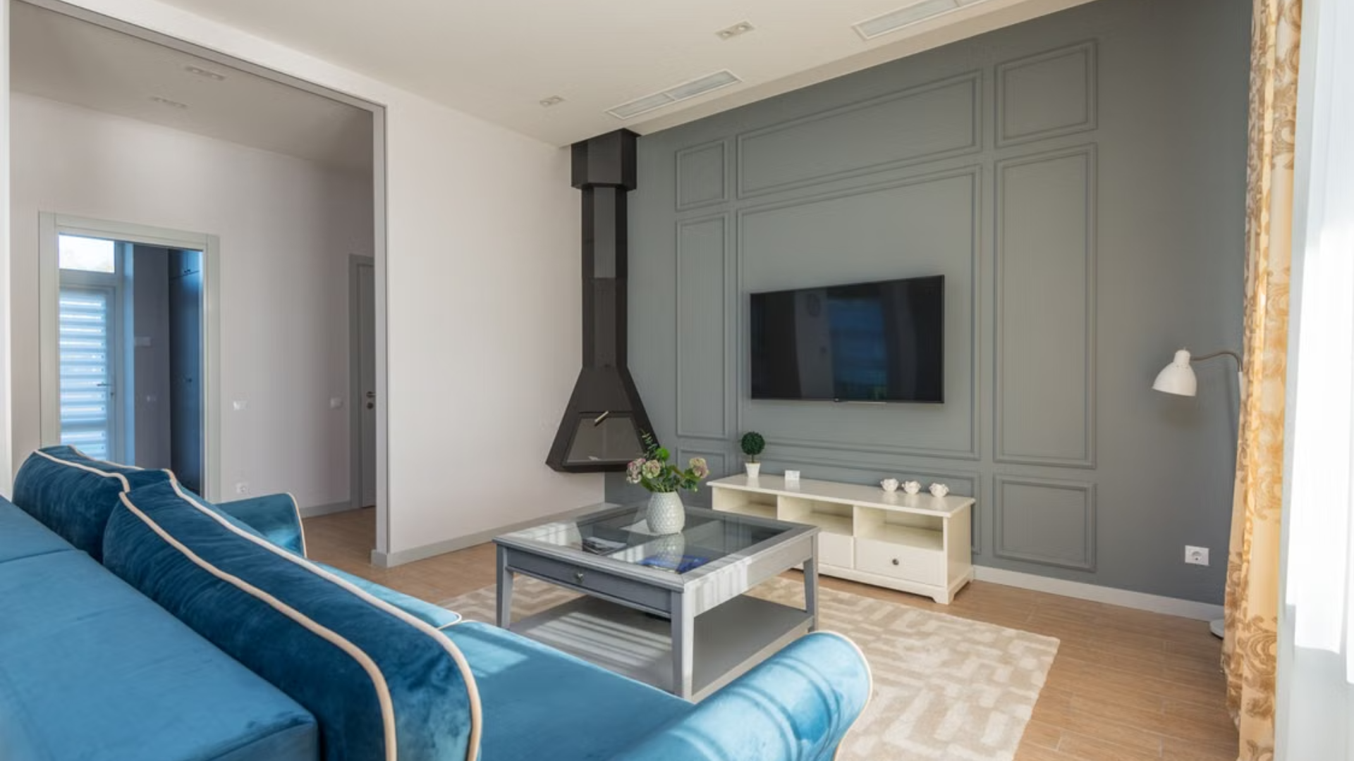 Modern living area featuring a bright blue velvet sofa, a TV mounted on a gray paneled wall, and a black corner fireplace.