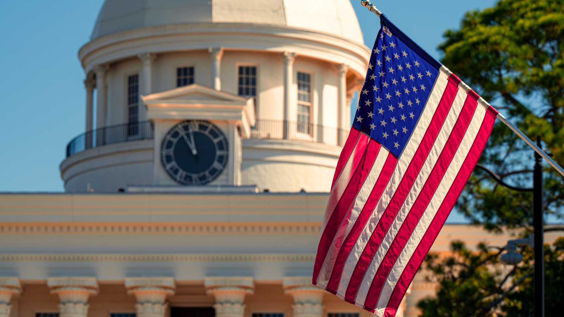 An American flag waving in the foreground, with the domed roof and clock of a white, classical-style building slightly out of focus behind it.