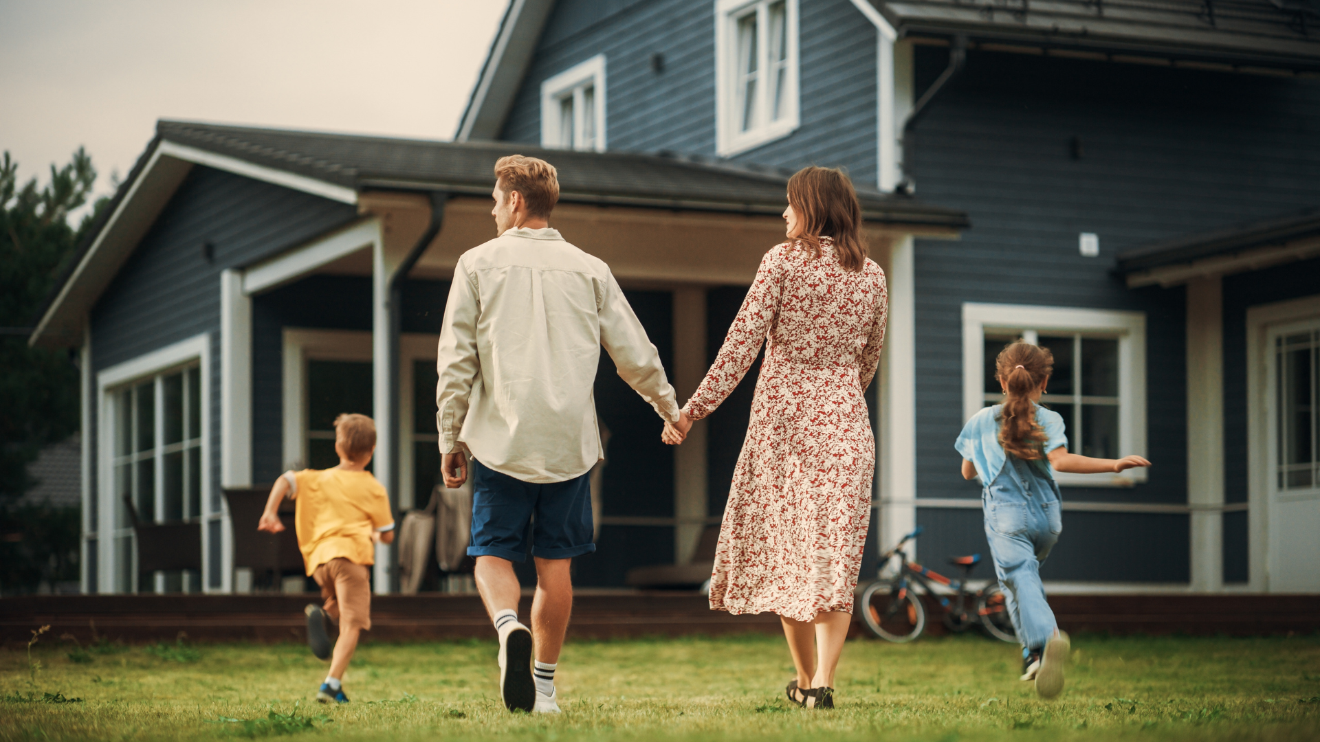 A family walking across a lawn towards a large gray house, with the parents holding hands and two children running ahead.