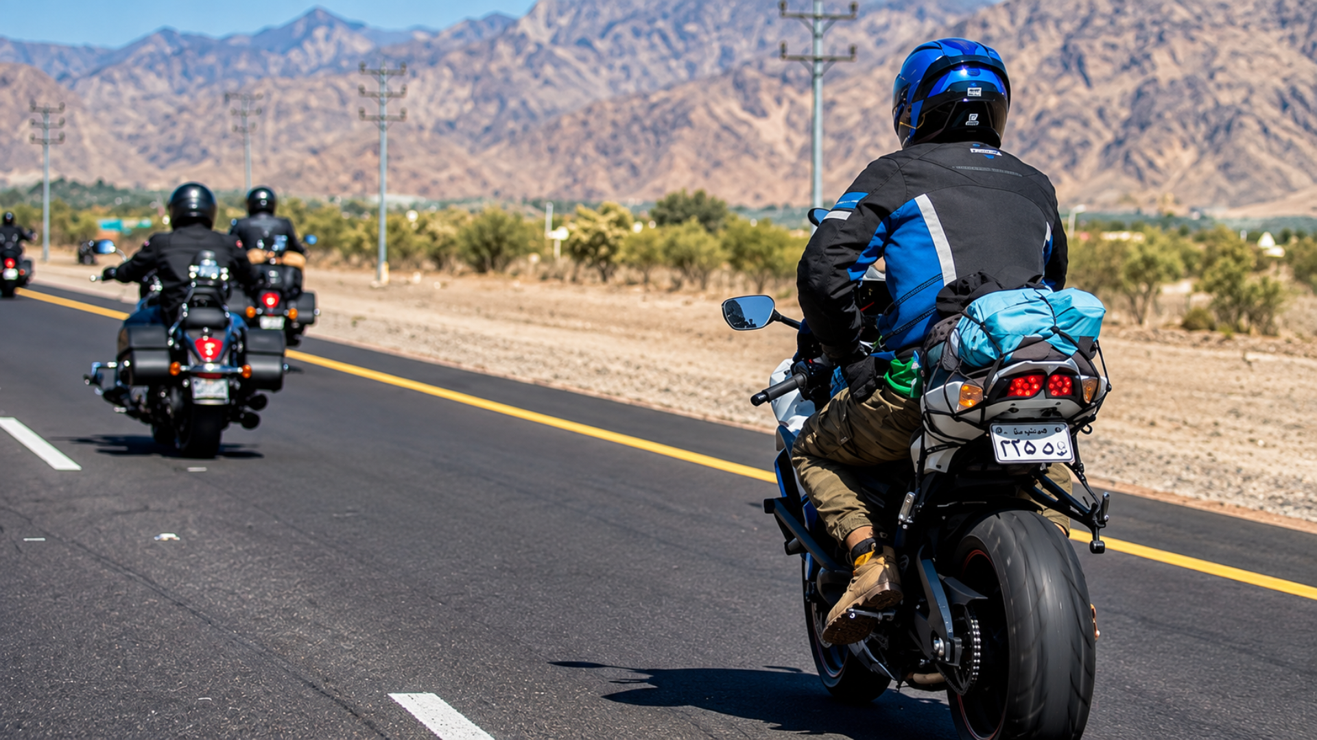 Rear view of a motorcyclist riding along a desert highway with mountain scenery in the background.