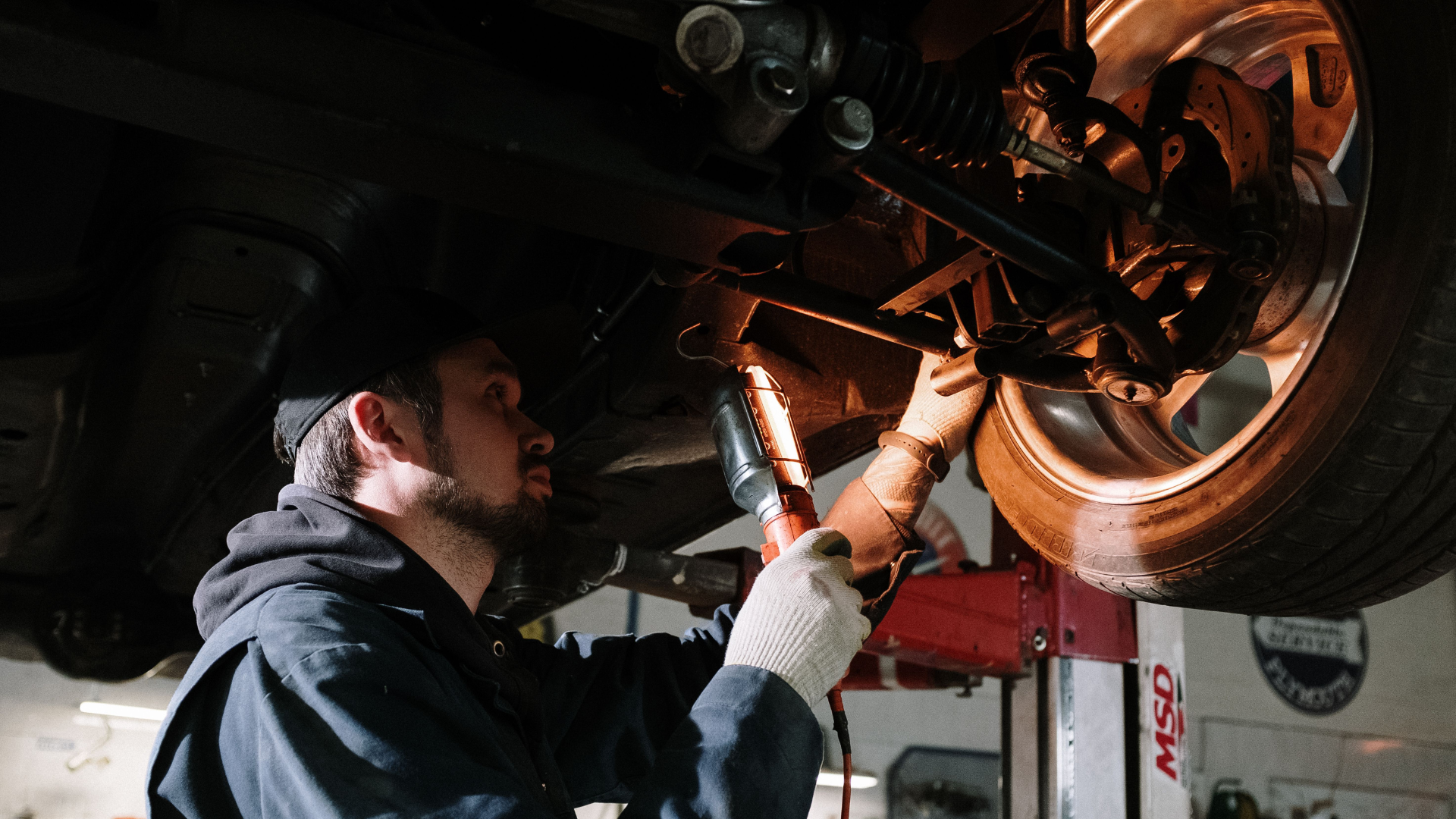 A mechanic holding a work light while inspecting the undercarriage and wheel assembly of a car.