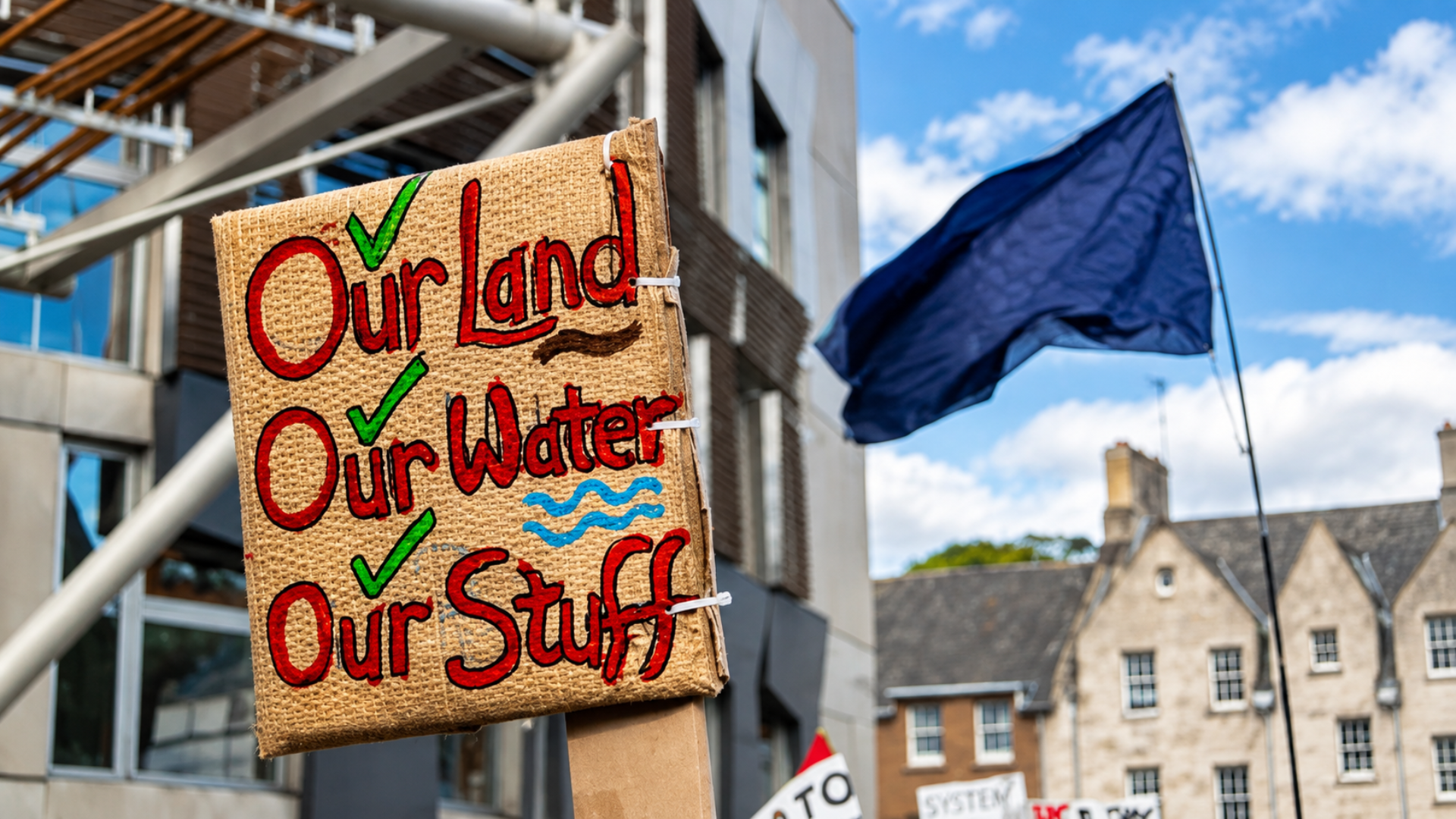 Burlap protest sign reading "Our Land, Our Water, Our Stuff" displayed next to a dark blue flag.