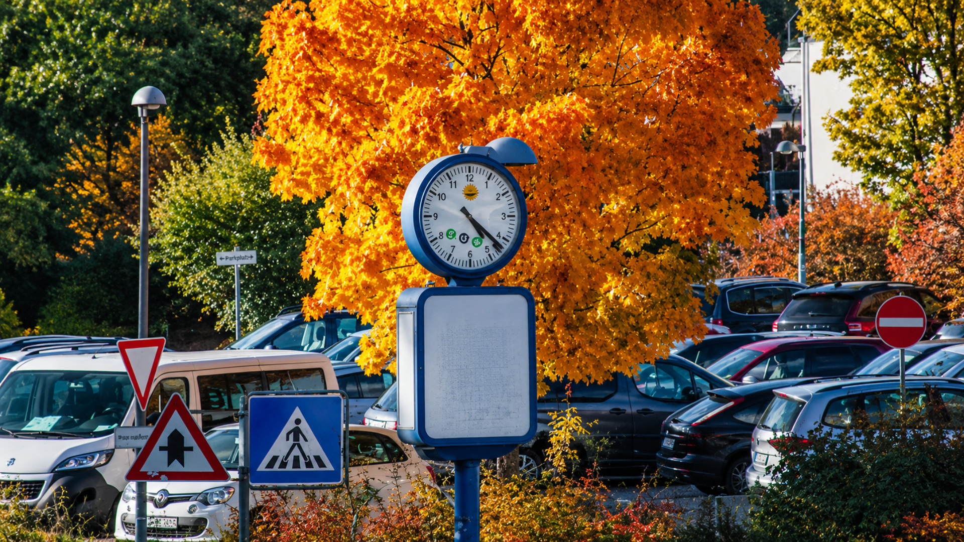 Outdoor public clock mounted on a post in a parking lot surrounded by autumn trees.
