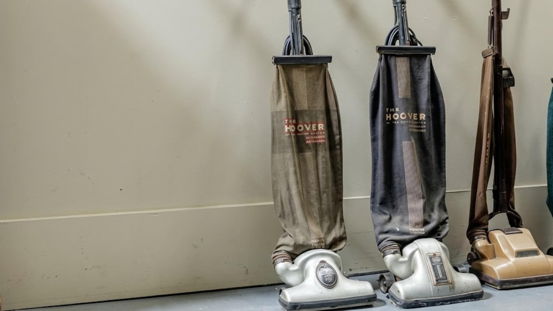 A row of three vintage vacuum cleaners stands against a wall, showcasing different models and colors.