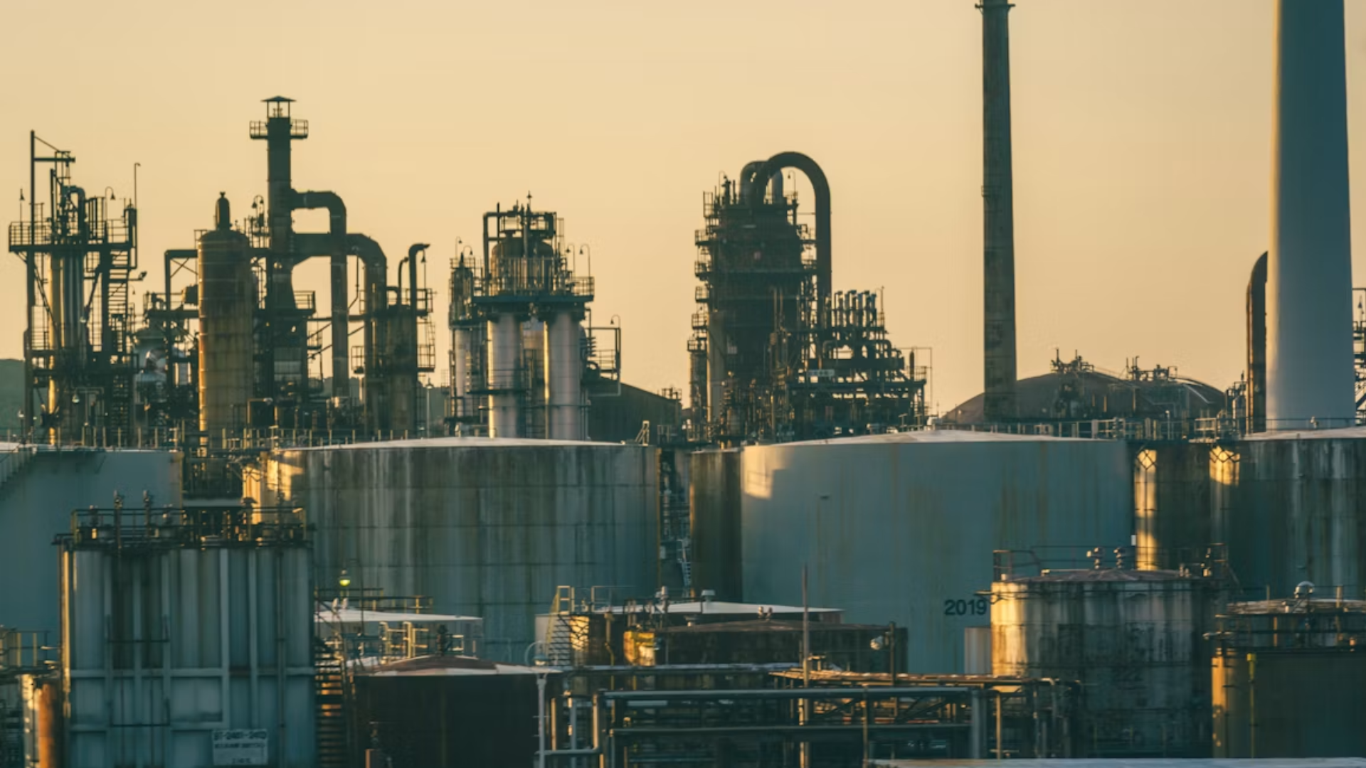 Industrial oil refinery at sunset, with towering distillation columns, storage tanks, pipes, and smokestacks silhouetted in warm golden light.