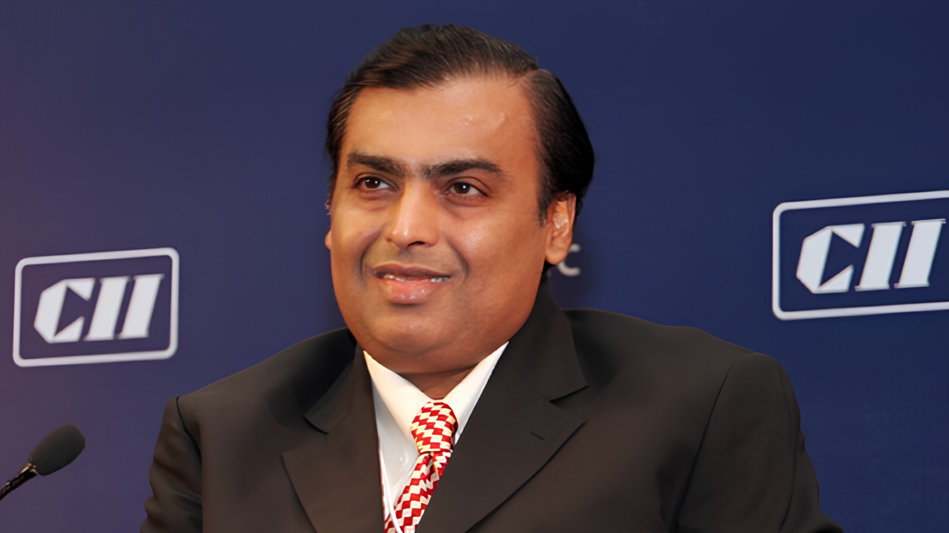 Indian billionaire Mukesh Ambani sits on a stage at a panel event in front of a blue backdrop.