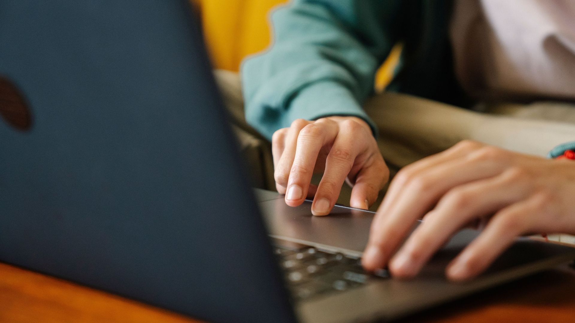 Close-up of a person in a green jacket typing on a laptop at a wooden table.