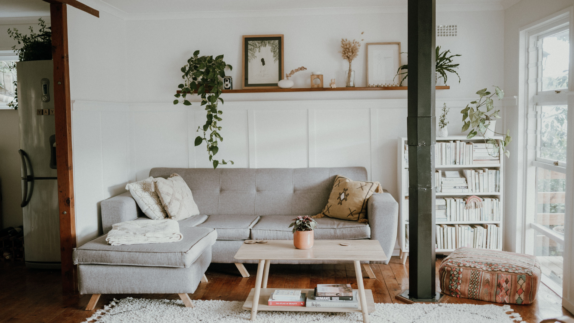  A brightly lit, bohemian-style living room features a light grey sectional sofa, a low wooden coffee table, potted houseplants, and a white bookshelf.