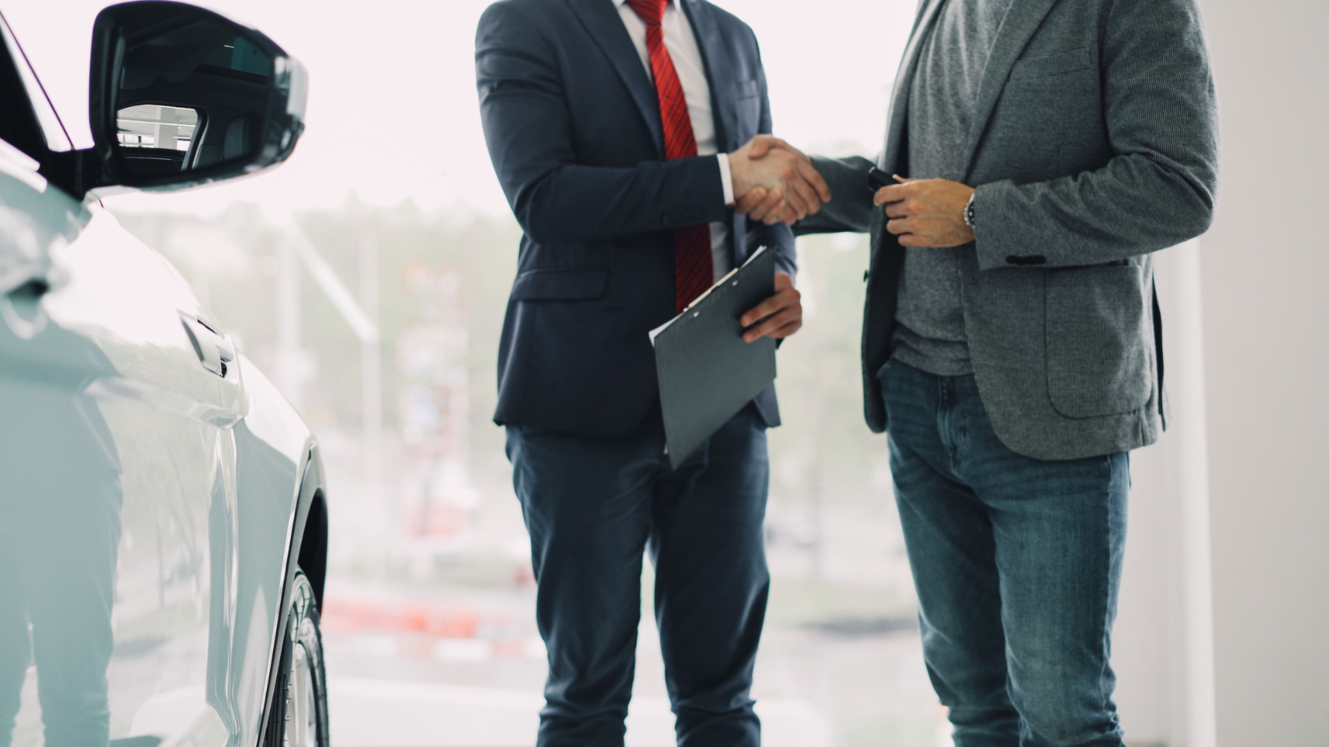 A car salesperson in a dark suit and red tie shakes hands with a customer next to a white vehicle.