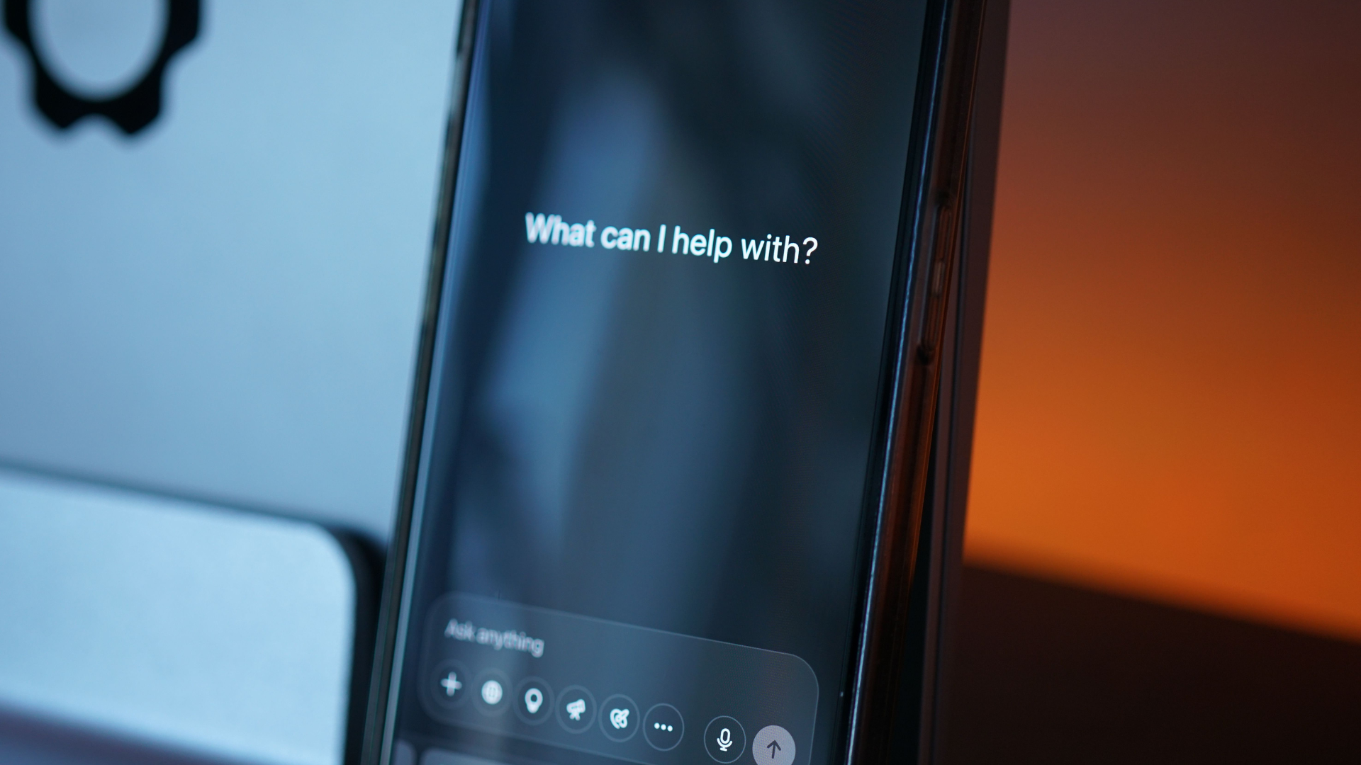 A close-up shot shows a smartphone displaying an AI app interface with the text _What can I help with__ propped up against a laptop.