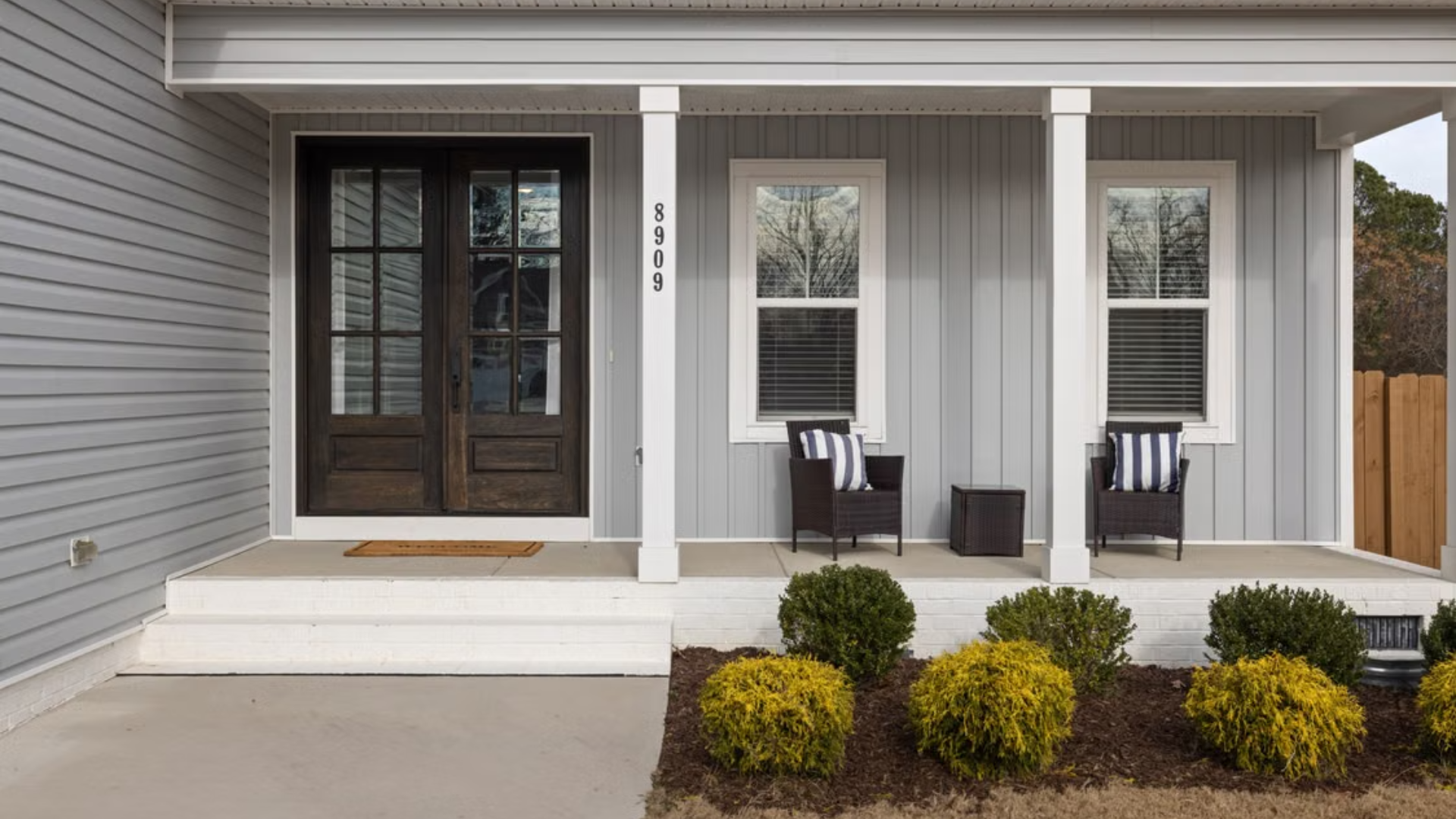 A cozy front porch on a light grey house is outfitted with dark wood double doors, two armchairs, and small garden shrubs.