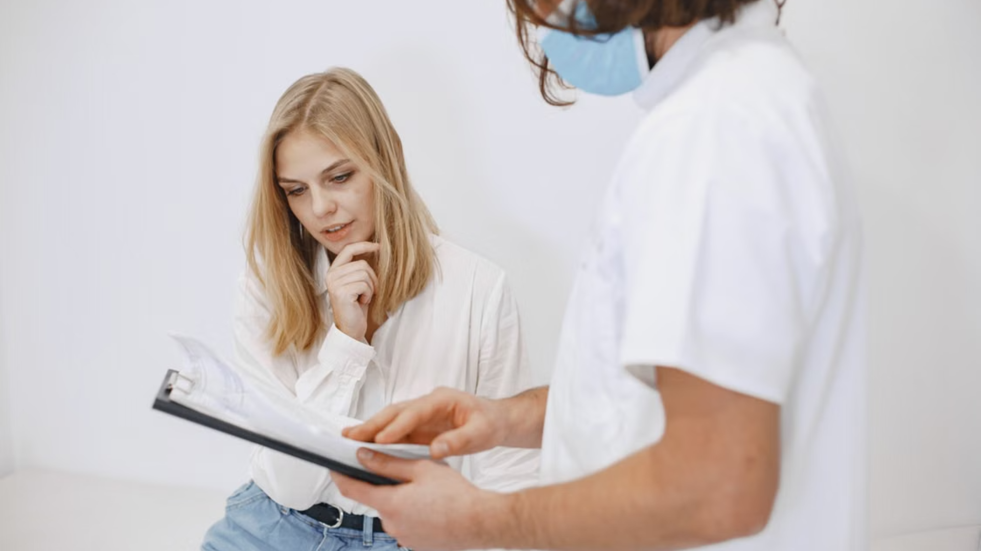  A healthcare professional wearing a face mask reviews medical paperwork on a clipboard with a blonde female patient.
