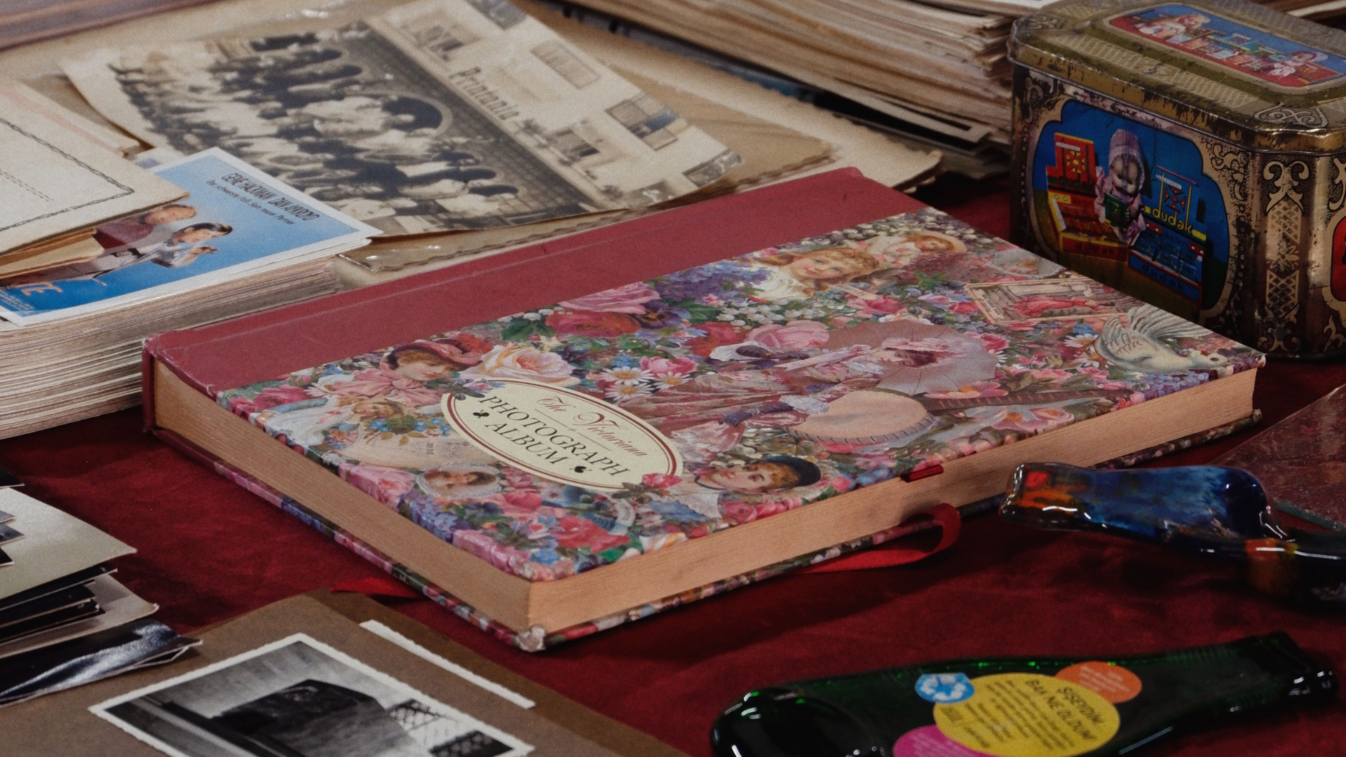 A high-angle close-up displays an array of vintage items, photo albums, loose black-and-white photographs, and old tins laid out on a table.