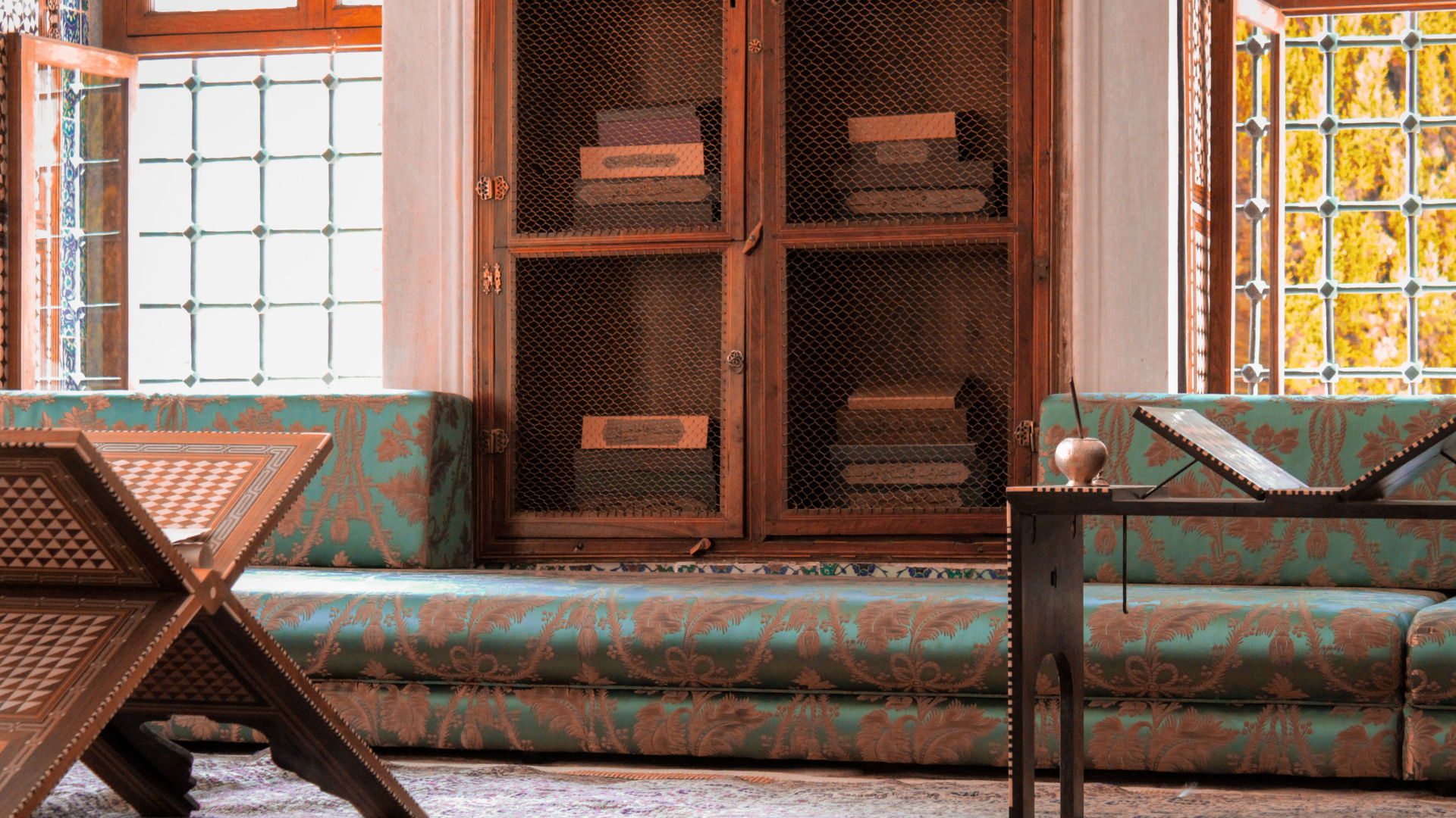 A low, bench-style seating area upholstered in teal floral fabric sits beneath a large mesh-front wooden book cabinet between decorative windows.