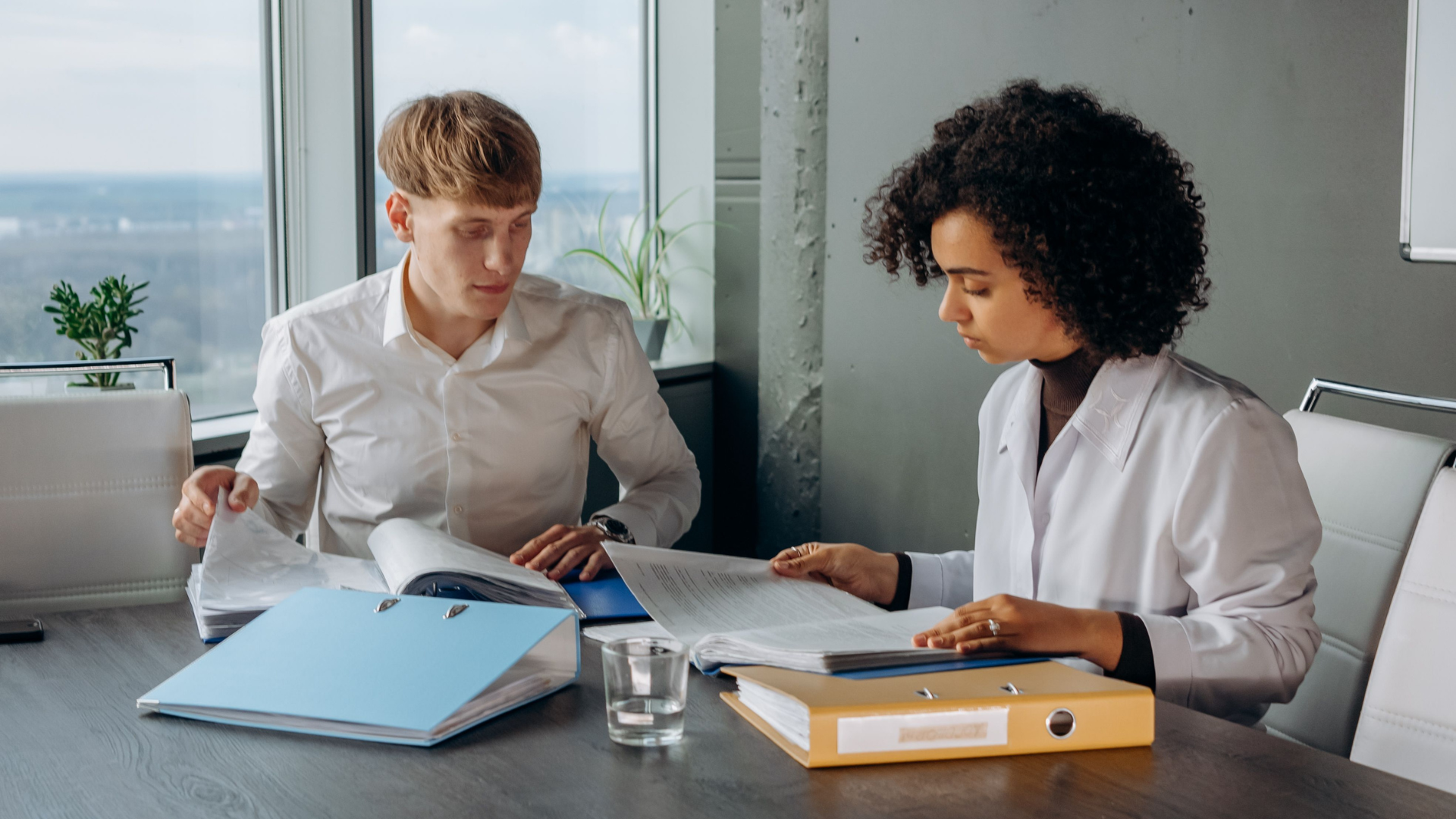 A man in a white dress shirt and a woman with curly hair seated at a conference table, both actively flipping through ring binders filled with paperwork.