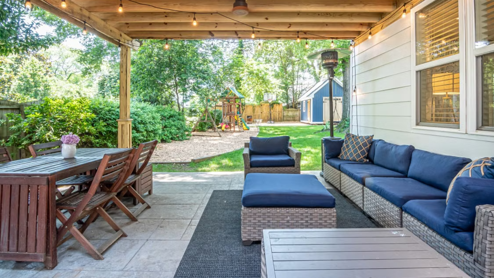 A spacious covered patio area is furnished with a wooden dining set, string lights, and a navy blue sectional sofa overlooking a backyard.