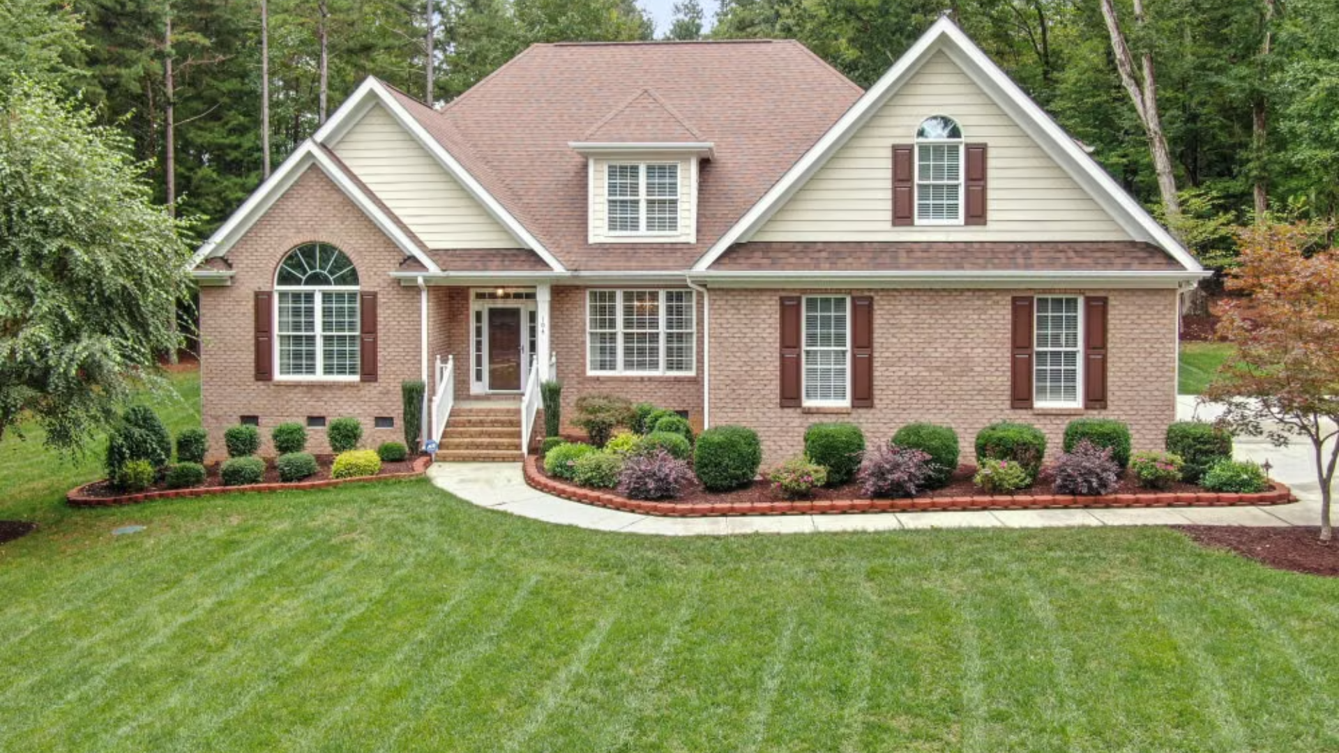 A two-story suburban brick house with beige siding features a manicured green lawn and a curved front garden bed.