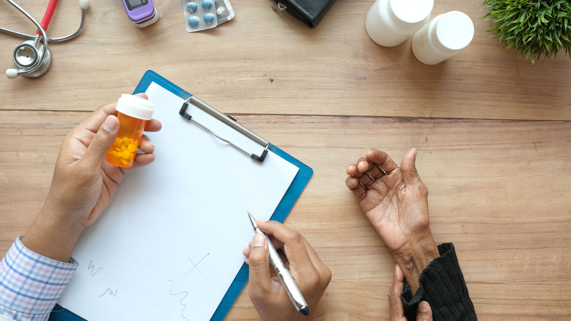 An overhead view shows a healthcare practitioner writing on a clipboard while holding a pill bottle next to a patient's hand on a wooden table.