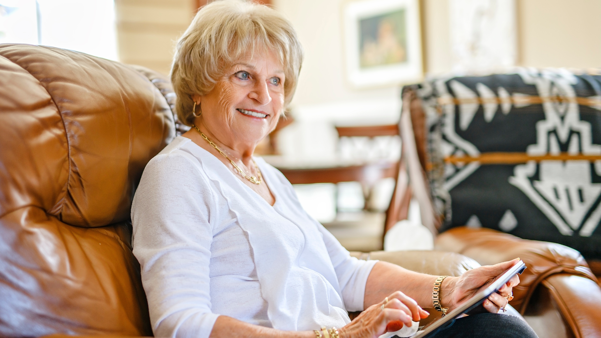 An older woman with short blonde hair sits comfortably in a brown leather armchair while holding a tablet and smiling.