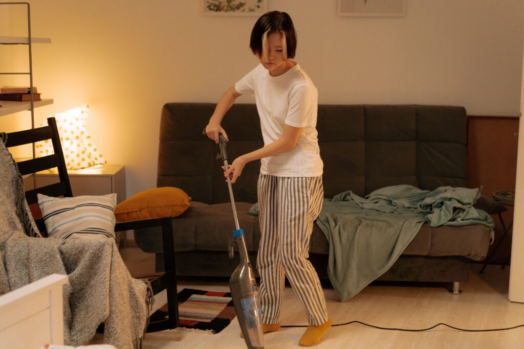 Person vacuuming a lived-in living room during an evening cleanup, with blankets and cushions still on the sofa.