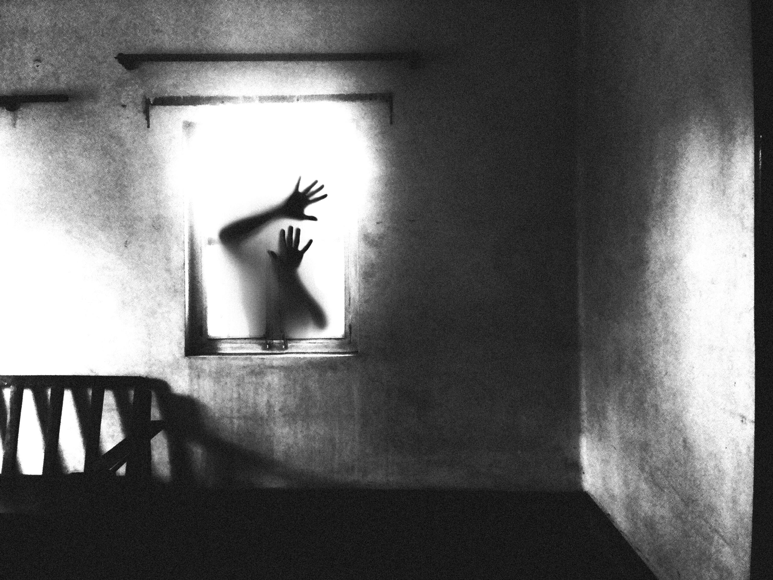 Shadowy silhouette with outstretched hands visible through a brightly backlit window in a dim interior room.