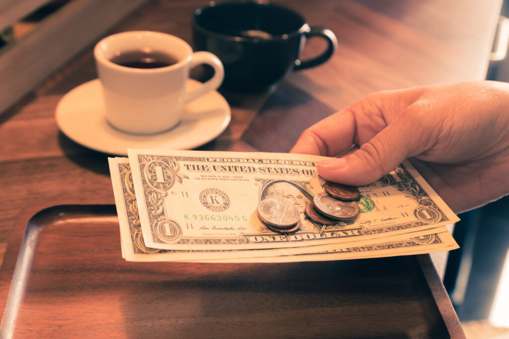 Person paying with dollar bills and coins beside cups of coffee.