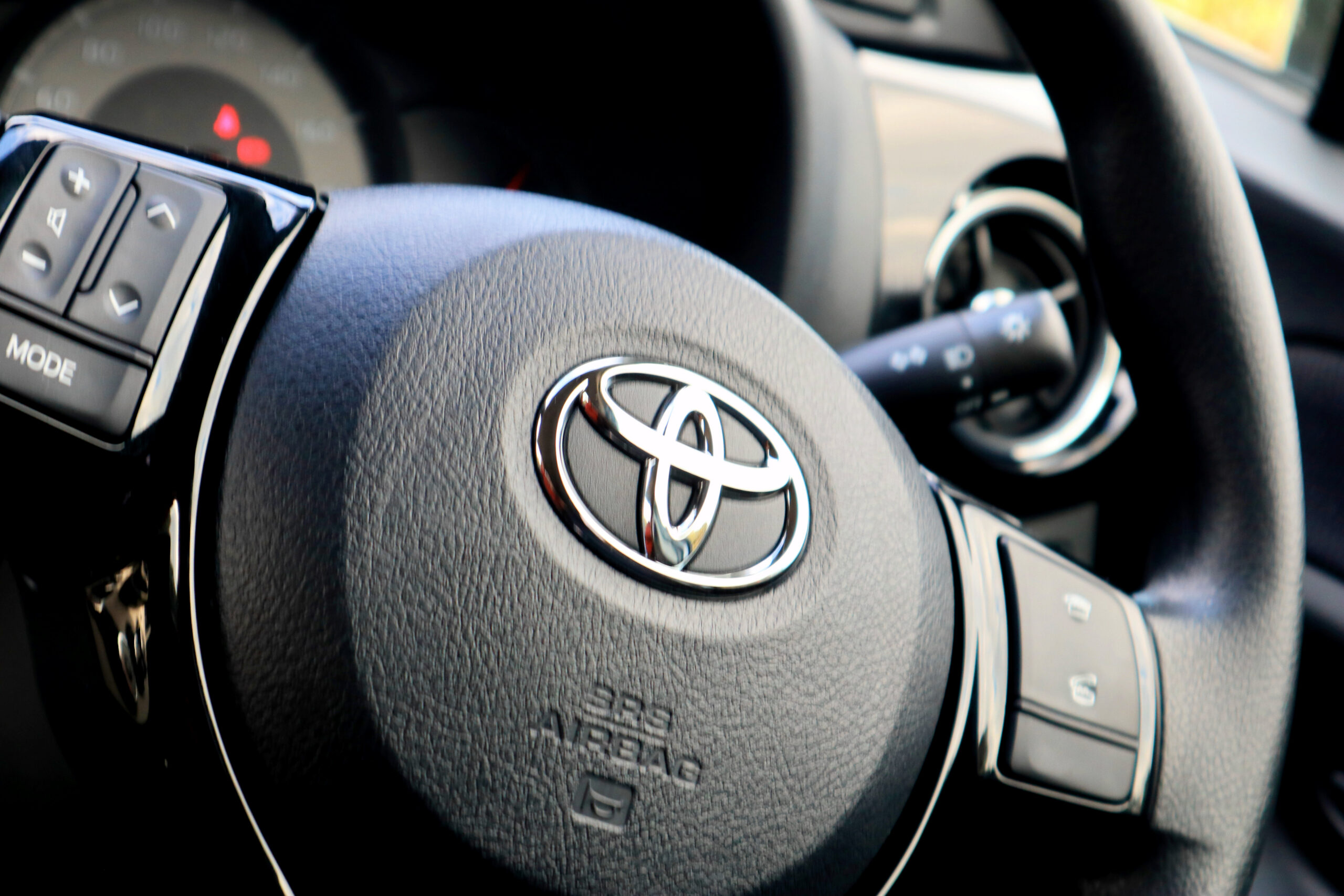 Close-up of a Toyota steering wheel with chrome logo, audio and mode controls, dashboard gauges, and ignition stalk in a modern car interior.