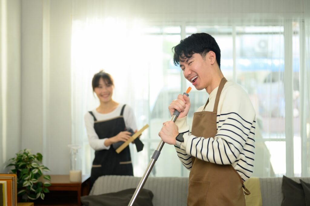 Smiling couple cleaning their bright living room together as one person mops the floor and the other holds a cleaning tool.