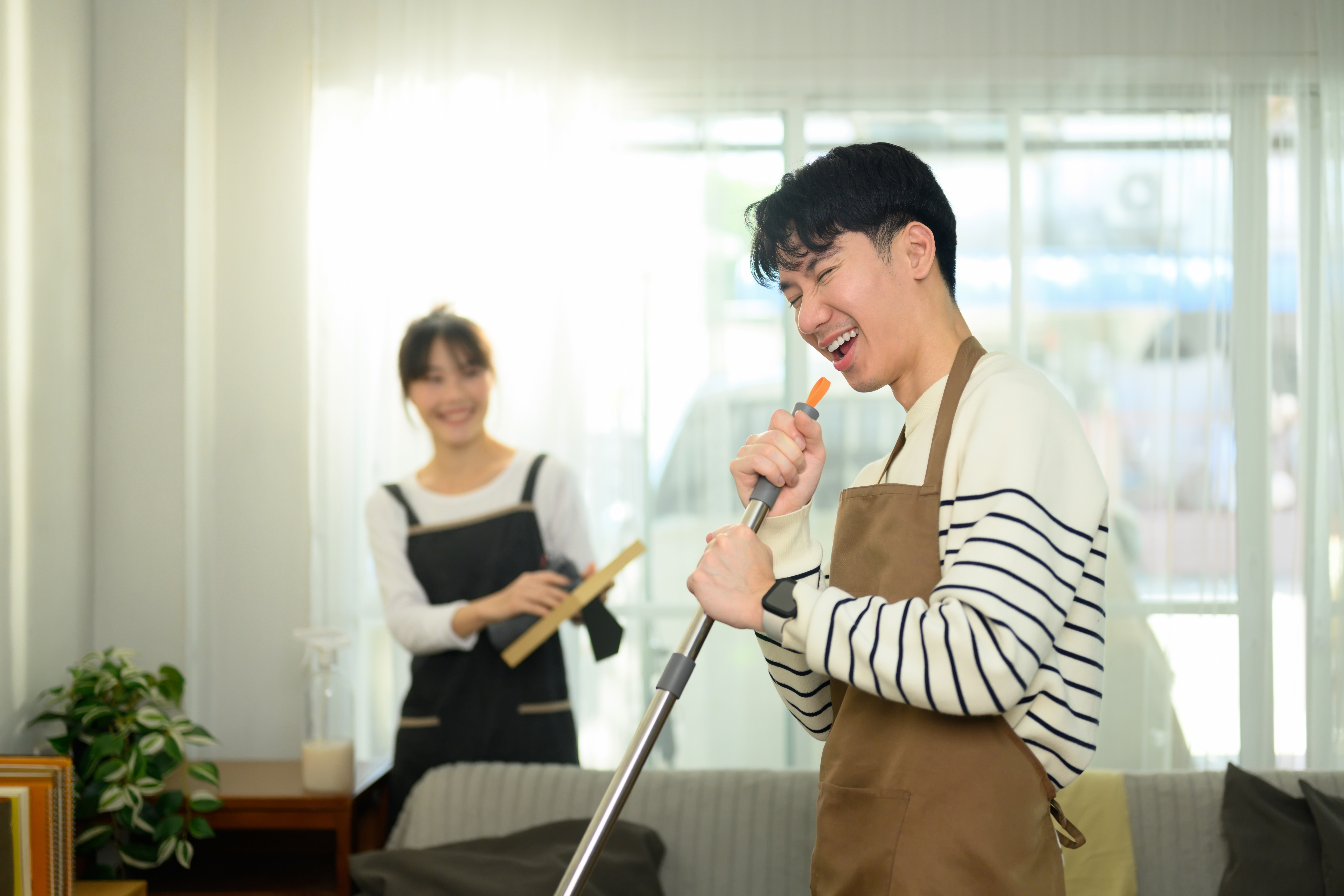 Smiling couple cleaning their bright living room together as one person mops the floor and the other holds a cleaning tool.