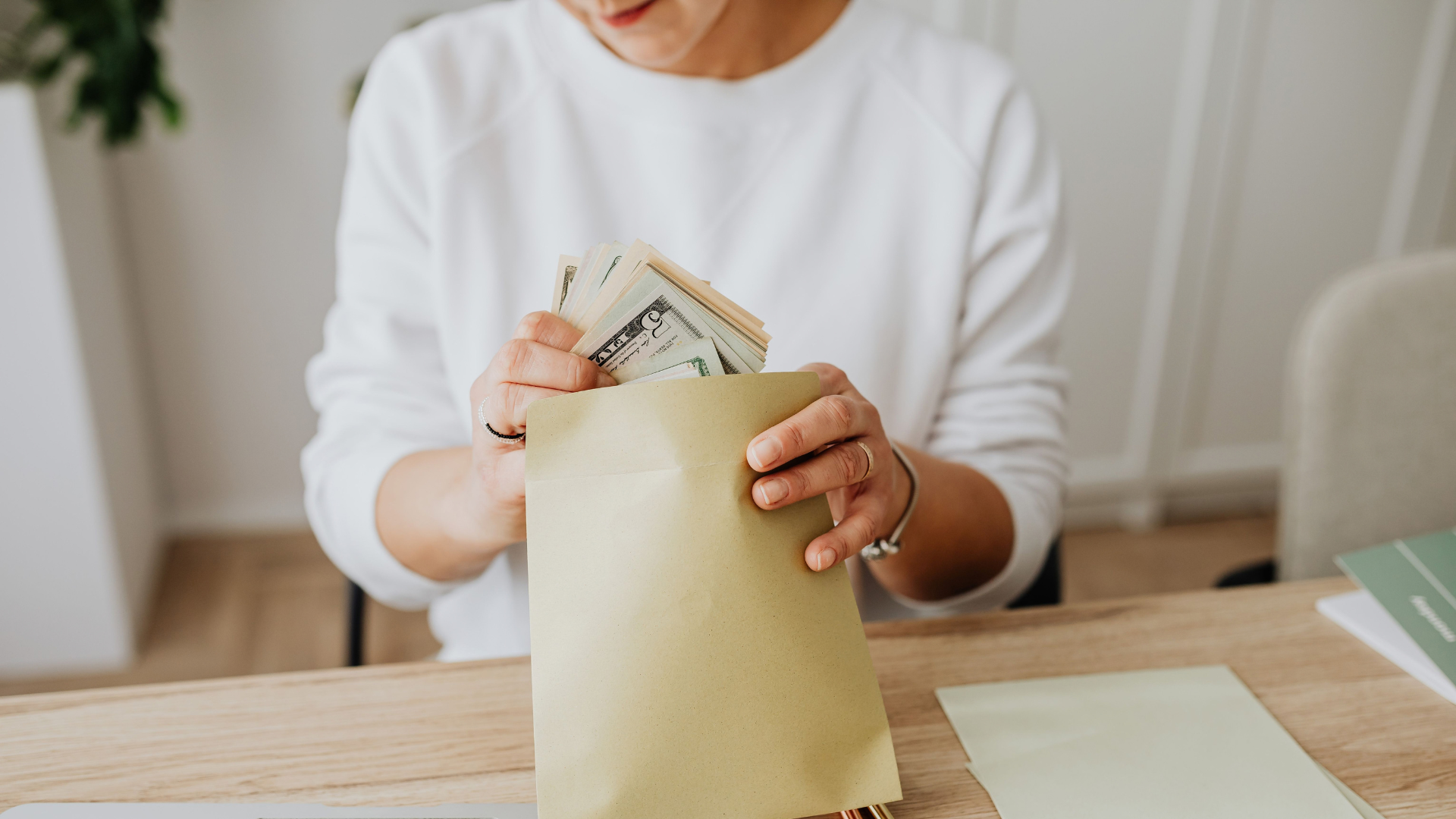 Close-up of person holding envelopes with cash at a wooden desk indoors.