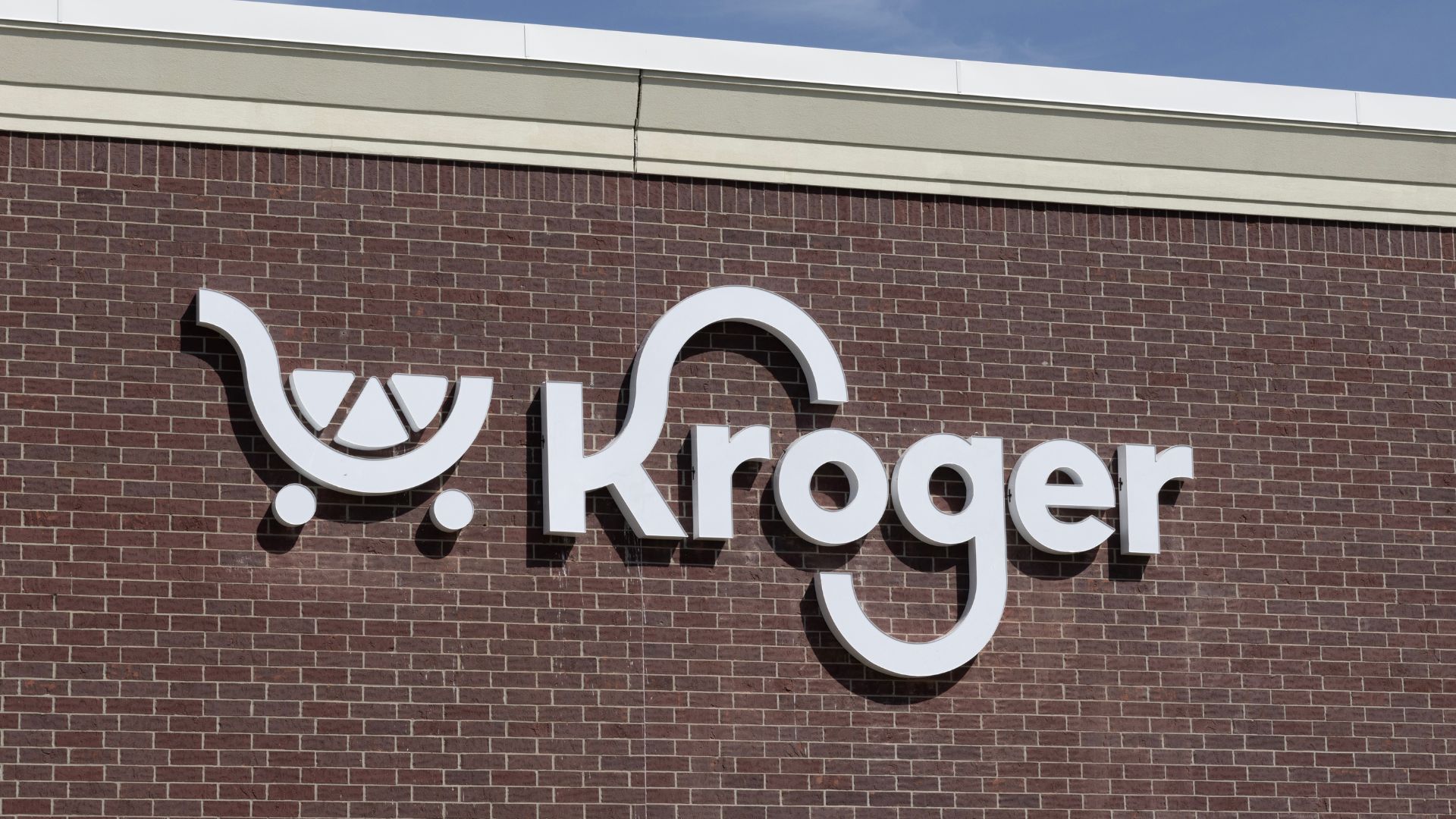 White Kroger supermarket logo and shopping cart symbol mounted on the exterior brick wall of a store.