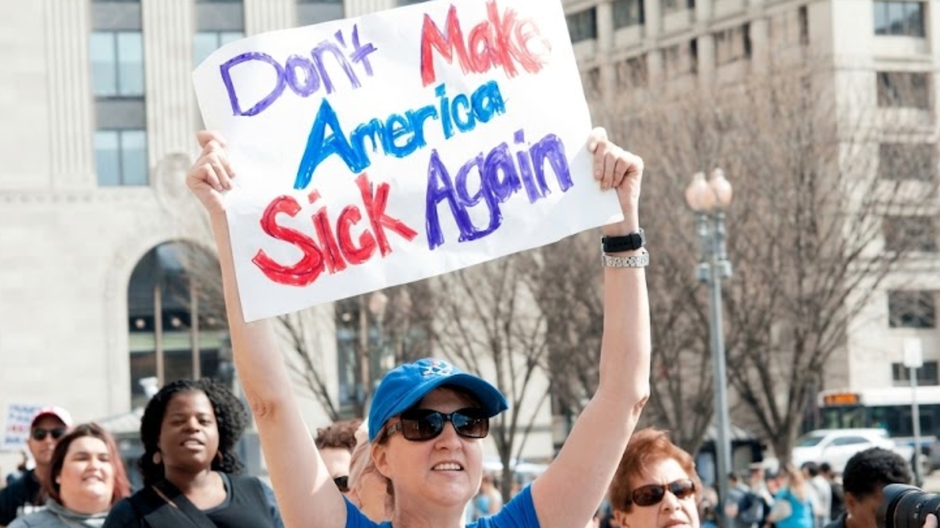 A crowd gathers at a public demonstration, with a woman holding a sign reading “Don’t Make America Sick Again.”