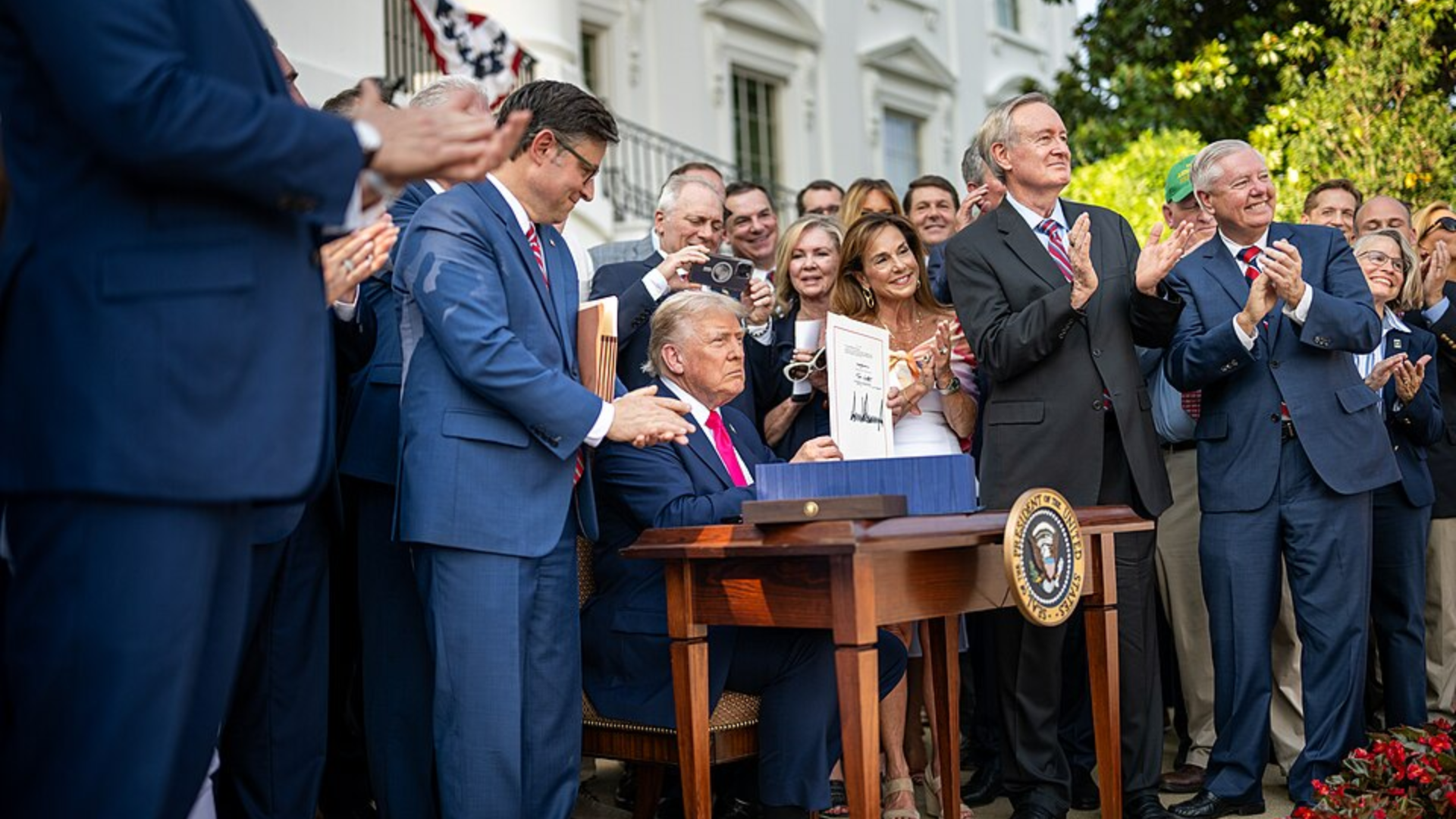 President Donald Trump holding up a signed document while surrounded by lawmakers at a White House event.