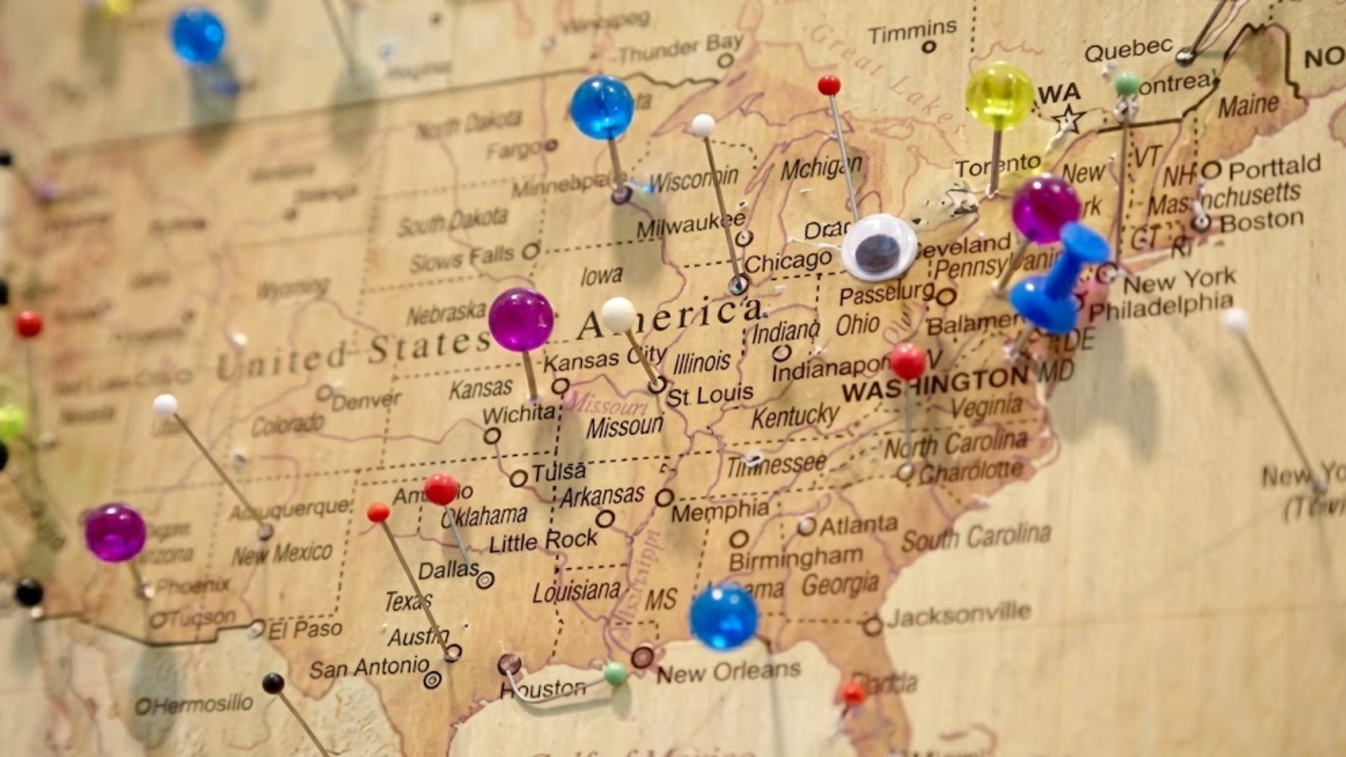 A wooden map of the United States marked with colorful push pins in various cities.